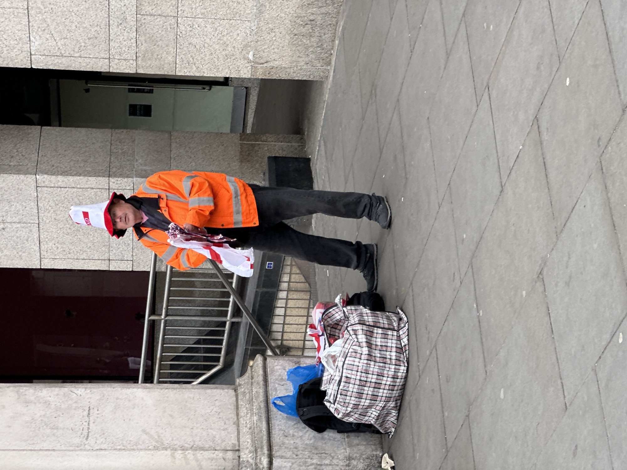 A man stands on a gray concrete sidewalk in an urban environment, wearing an orange safety jacket, black trousers, and black shoes. He is also wearing a white and red bucket hat. He appears to be selling small flags with the St. George's Cross. Next to him is a plaid bag filled with similar flags. The background includes a building facade with a recessed doorway and metal railings. The location is London, United Kingdom.