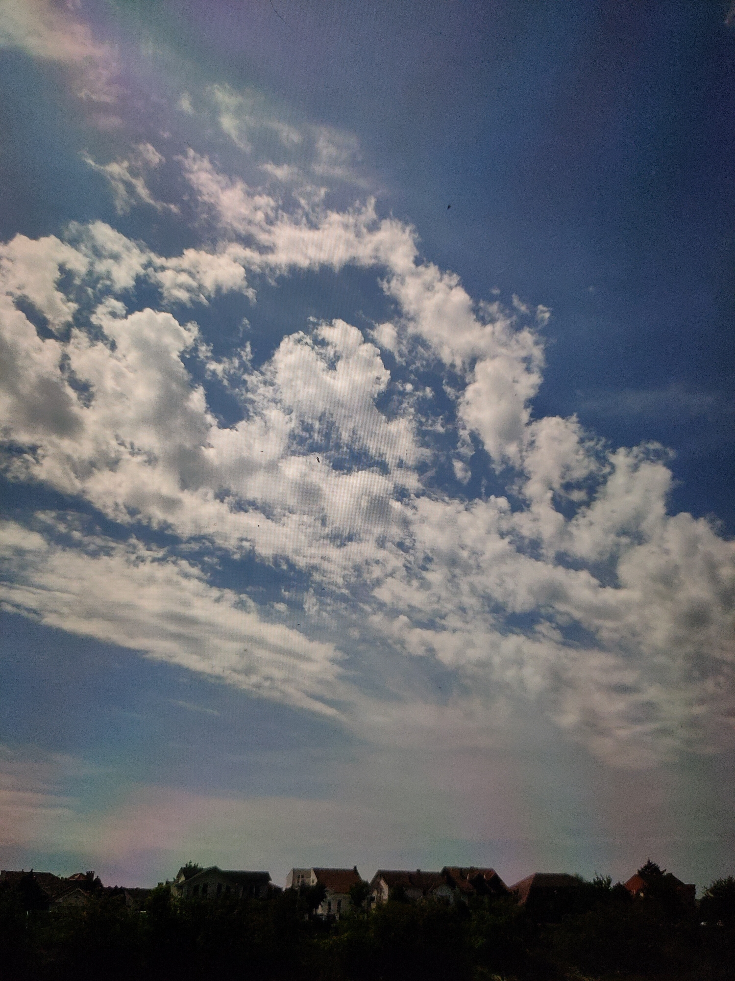 An eye-level, outdoor, medium shot captures a bright blue sky filled with scattered white and gray clouds. A thin band of rainbow-like colors is visible in the sky, toward the left side of the frame. Below the sky, a silhouette of a tree line is visible, dotted with the rooftops of several houses. The roofs are a mix of red and brown tiles, and the houses appear to be of various sizes and styles. The overall lighting suggests it is daytime, possibly late afternoon, given the diffused light and the colorful sky. The image has a slightly grainy texture, indicating it may have been taken with a digital camera.