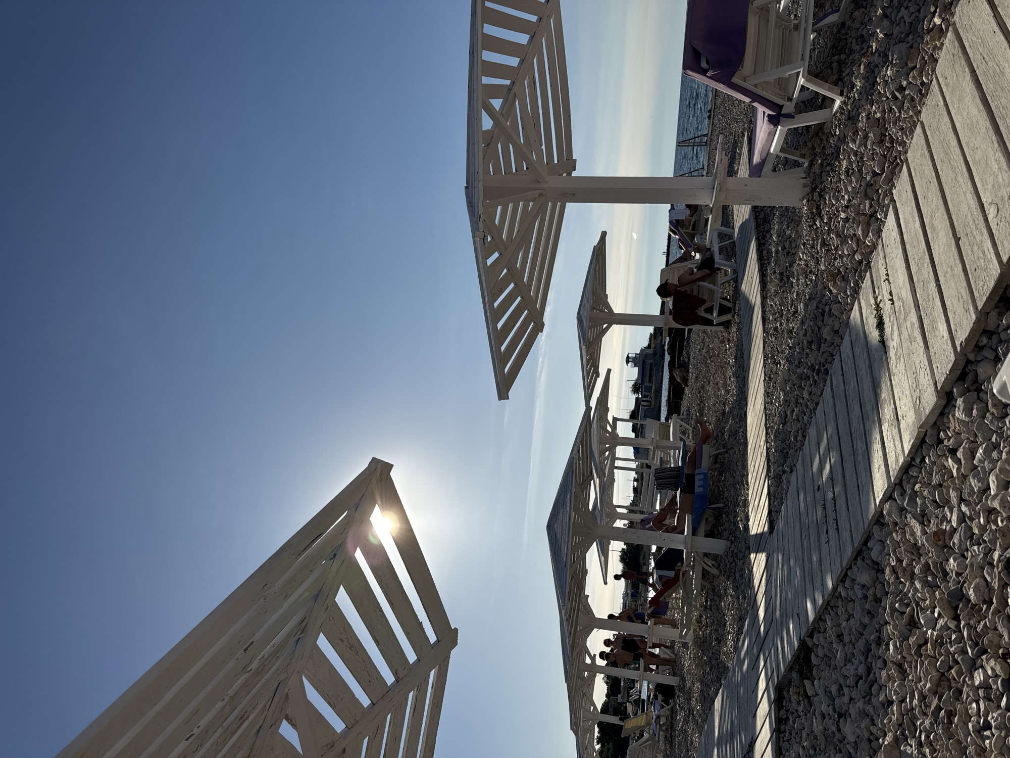 The image depicts a beach scene under a clear blue sky. In the foreground, a wooden boardwalk made of planks runs diagonally across a pebble-covered beach. Several white, slatted beach umbrellas are set up on the beach, providing shade. Sun loungers are placed beneath some of the umbrellas, with a few people visible relaxing on them. In the background, the sea stretches to the horizon.
The scene appears to be a beach resort or public beach area. The presence of beach umbrellas and loungers suggests a place designed for leisure and relaxation. The wooden boardwalk is typical for beach environments, providing a path over sand or pebbles. The overall atmosphere is one of a sunny day at the coast. Given the context of Sevastopol, Ukraine, this could be a popular tourist spot or a local beach within the city's limits.