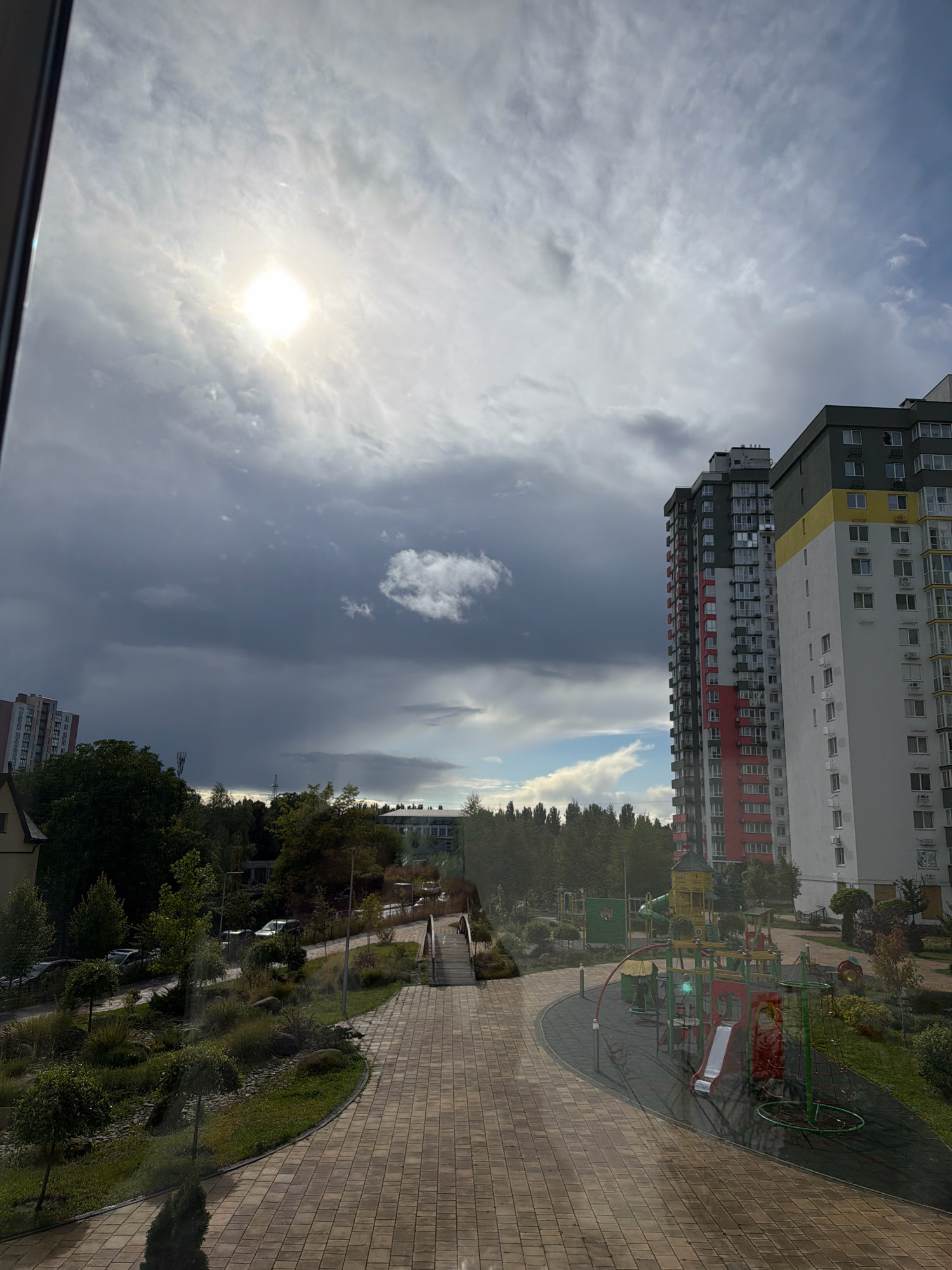 The image captures a view of a modern residential complex in Kyiv, Ukraine, under a dramatic, cloudy sky. The sun attempts to break through the heavy clouds, creating a bright, diffused light. In the foreground, a paved walkway leads through a landscaped area with lush greenery and decorative stones. Further on, a children's playground with colorful equipment, including slides and climbing structures, is visible. To the right, two tall, multi-story apartment buildings dominate the skyline. One building features a striking red and white facade, while the adjacent one is primarily white with yellow accents. The surrounding area is dotted with trees, and cars are parked along the road. The overall scene depicts a contemporary urban living environment with recreational spaces.