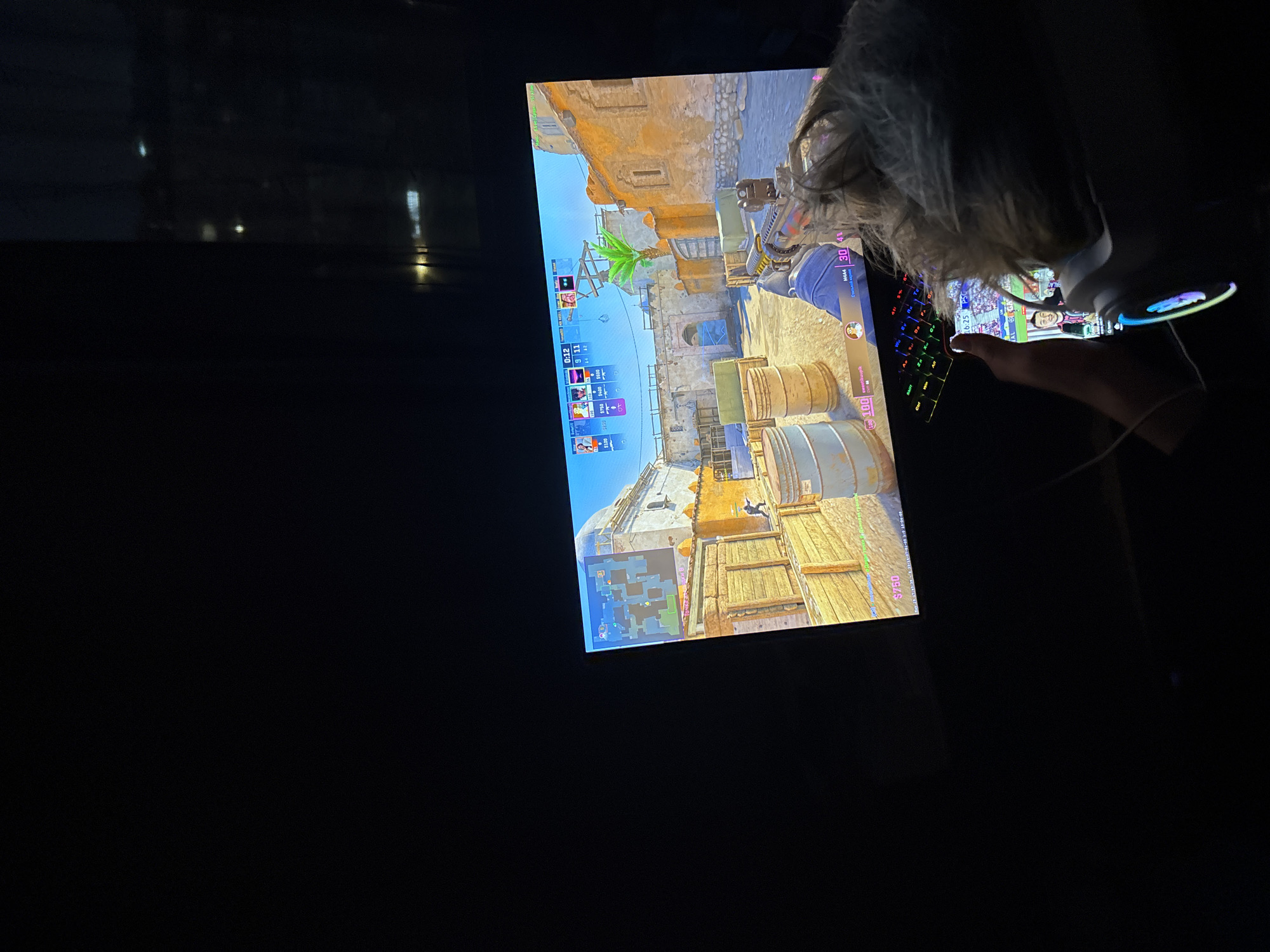 A person is playing the video game Counter-Strike: Global Offensive on a monitor in a dimly lit room. The game appears to be set on the map "Dust II" based on the environmental assets like the light tan buildings and wooden boxes. The person is using a keyboard and mouse to play the game. The player's perspective appears to be from the T side spawn on the map.