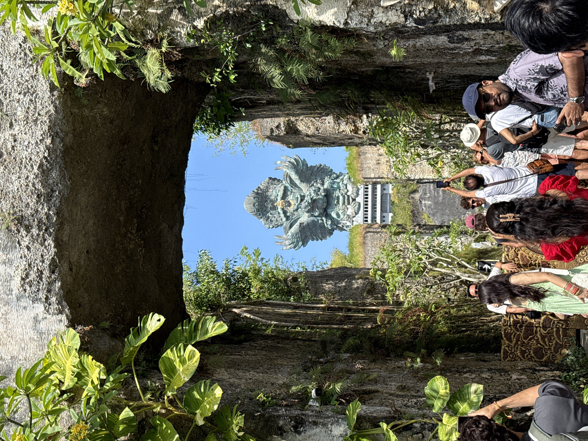 The image captures a view in Kuta Selatan, Indonesia, looking through a stone archway overgrown with vegetation. The archway frames a massive statue depicting a figure with wings, presumably the Garuda Wisnu Kencana statue. A building is seen behind the statue. To the right of the archway, a crowd of people are visible, some taking photos with their phones. The photo appears to be taken at eye level, looking upwards towards the statue.