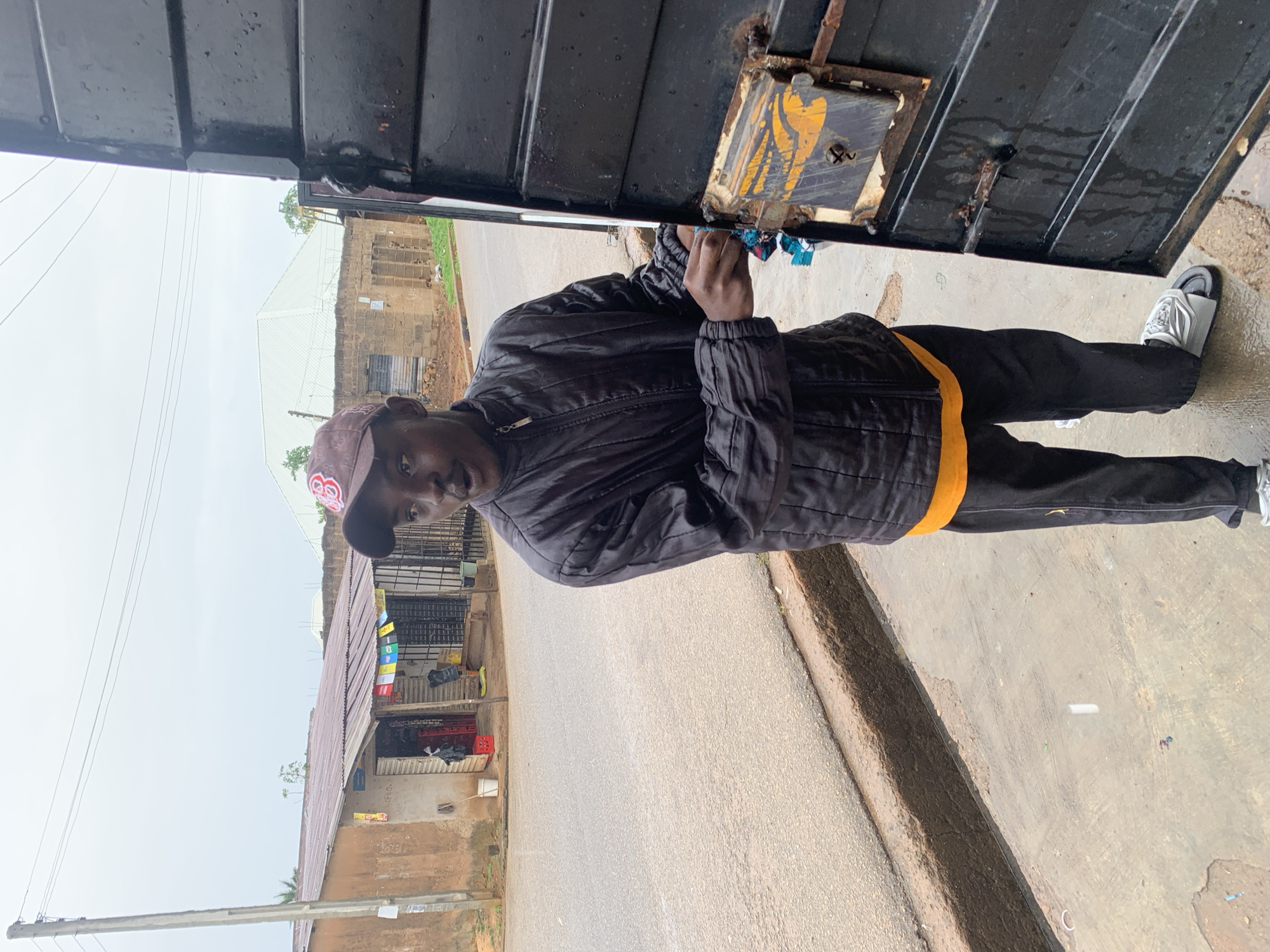 A young Black male wearing a black puffer jacket, an orange t-shirt, black pants, a brown baseball cap, and silver slip-on shoes stands next to an open black metal gate. He is looking to his right, towards the viewer's left, with his mouth slightly open. His right hand is near the latch of the gate, which is rusted and has yellow markings. The gate is partially open, revealing a dark interior.
Behind him, to the left, is a street with a raised concrete curb. On the other side of the street are several buildings, including a small shop with a corrugated metal roof. The shop has various items displayed in front of it. A power pole with wires runs vertically on the left side of the image. The ground appears wet, suggesting it may have rained recently. In the background, there are more buildings and a large white-tiled roof structure. The overall impression is an outdoor scene in a modest urban or semi-urban environment.