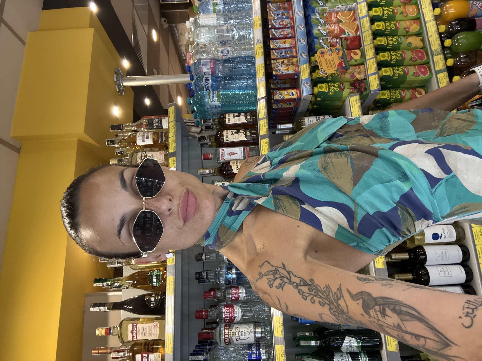 The image shows a woman with dark hair and sunglasses, wearing a colorful, patterned dress. She is taking a selfie in a well-lit store, likely a supermarket or liquor store, with aisles of beverages visible behind her. The shelves are stocked with various bottles of alcohol, including vodka, rum, and wine, as well as bottled water and juice cartons.
Notable text visible on the products includes brand names like "Smirnoff," "Finlandia," "Beefeater," "Black Ram," "Highland Queen," and "Devin" water. Juice brands like "Happy Day" and "Brauch" are also present. Some Cyrillic text is also visible on several labels.
The woman has a relaxed expression and is looking at the camera. The lighting suggests it's daytime, and the indoor setting indicates she is shopping. The background is filled with products, creating a commercial and consumerist atmosphere. The overall scene is a snapshot of a person in a shopping environment, focusing on her and the surrounding merchandise. The orientation of the image is such that the woman appears to be standing upright, but the rest of the scene is rotated.