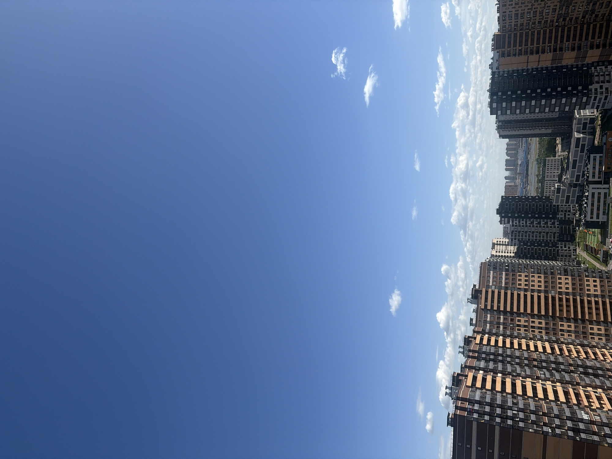 The image shows a view of a cityscape under a blue sky with scattered clouds. The buildings are high-rise residential structures with a mix of brown and black exteriors. The architecture is modern, with many windows visible. The scene appears to be in Novosaratovka, Russia. There are no people visible in the image. The environment suggests a clear day.