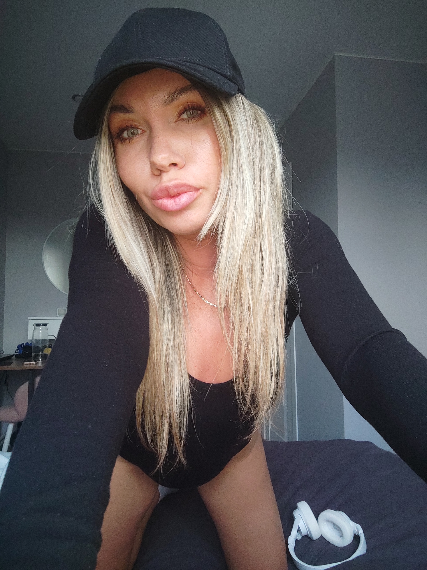 A young woman is depicted in a close-up, chest-up shot, appearing to be taking a selfie indoors in Votkinsk, Russia. She has long, straight blonde hair and light-colored eyes, possibly green or hazel. Her lips are full, and she has a slightly pouting expression. She is wearing a black baseball cap and a form-fitting, long-sleeved black bodysuit or top, accessorized with a delicate, thin gold-colored chain necklace.
The woman is positioned on what appears to be a bed with dark grey or blue bedding, with white over-ear headphones resting on the bed to her right. The background features plain, dark grey walls, suggesting a modern interior space, possibly a bedroom or living area. On the left side of the frame, behind her, a table is visible with a round mirror on the wall above it, and a glass jar or bottle along with other indistinct items on the table surface. The lighting suggests it is daytime, with soft natural light illuminating the scene. There is no visible text in the image.