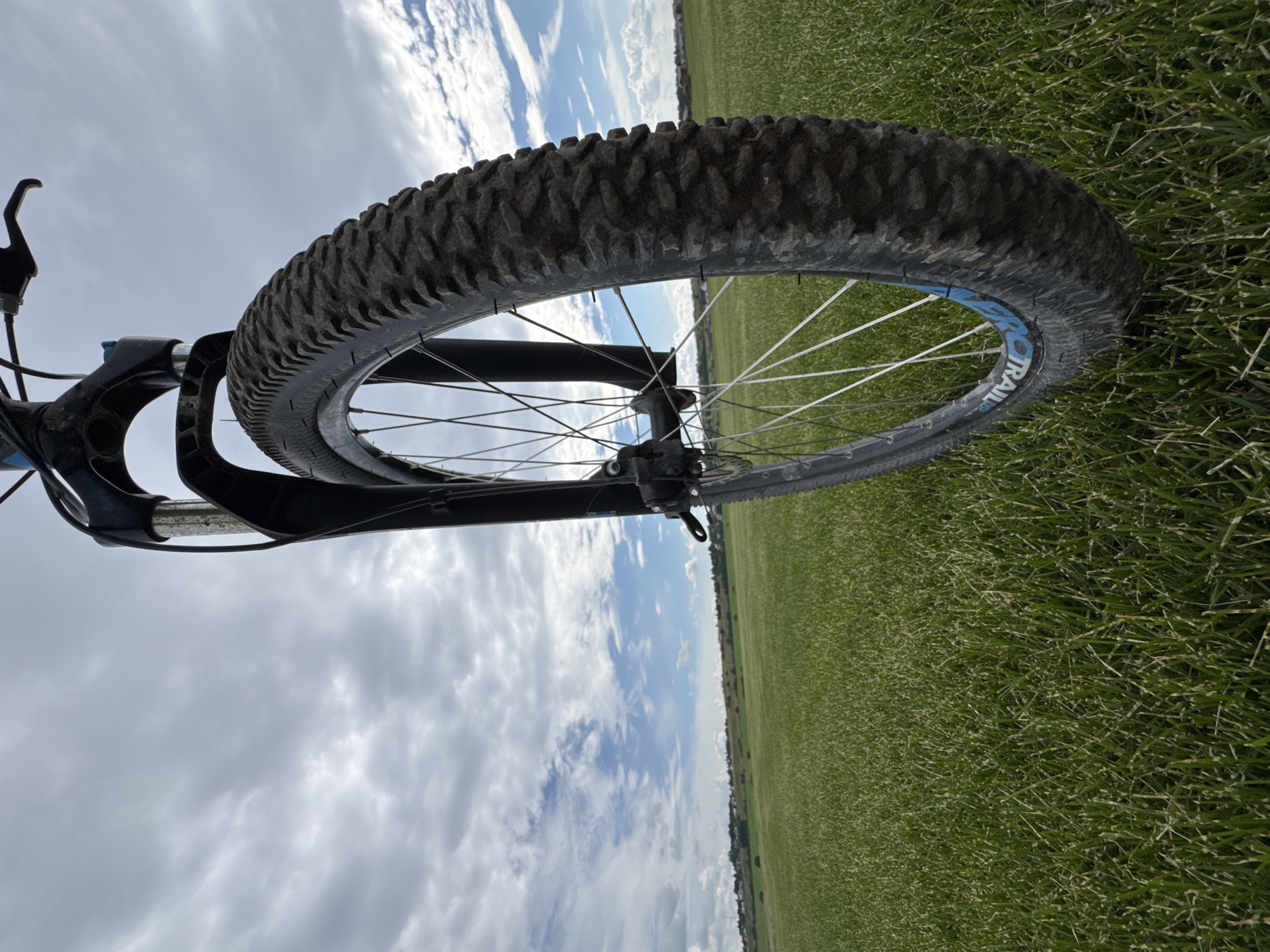 The image shows a close-up, low-angle view of a bicycle's front wheel and forks, positioned in a grassy field under a cloudy sky. The bicycle appears to be on a trail or open area, with a treeline and some scattered buildings visible in the distance on a gentle hill. The foreground is dominated by lush green grass, with the knobby tread of the bicycle tire clearly visible. The sky is filled with dramatic, textured clouds, with hints of blue sky peeking through. The location is identified as Kempelevo, Russia.