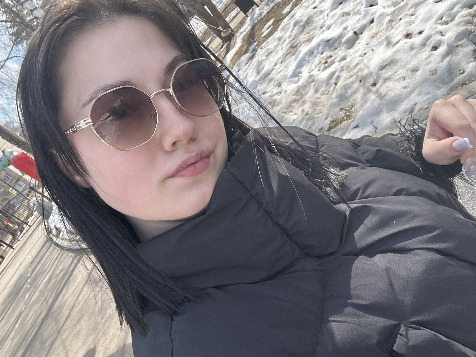 The image features a young person, likely female, taking a selfie outdoors in what appears to be a cold, bright, late winter or early spring setting in Chaikovsky, Russia. The subject is positioned in the foreground, angled slightly, looking towards the camera with a neutral to slightly serious expression.
She is wearing stylish, hexagonal-framed sunglasses with gold-colored frames and dark lenses, which obscure her eyes. Her dark, straight hair is parted down the middle and falls around her face and shoulders. She is dressed in a thick, black puffer jacket, indicating cold weather. A hint of dark faux fur trim is visible near her shoulder, possibly part of the jacket's collar or hood. Her fingernails are manicured with light-colored polish, possibly a French tip style.
The background shows patches of melting snow covering the ground, interspersed with bare earth. Strong, direct sunlight illuminates the scene, casting clear shadows on the ground, indicating it is daytime. Bare tree branches are visible against a bright sky. In the distance, parts of what appear to be urban or residential buildings can be vaguely seen, including what looks like a fire escape on one structure in the left background. The overall scene suggests an urban or semi-urban environment in a cold climate. There is no visible text in the image.