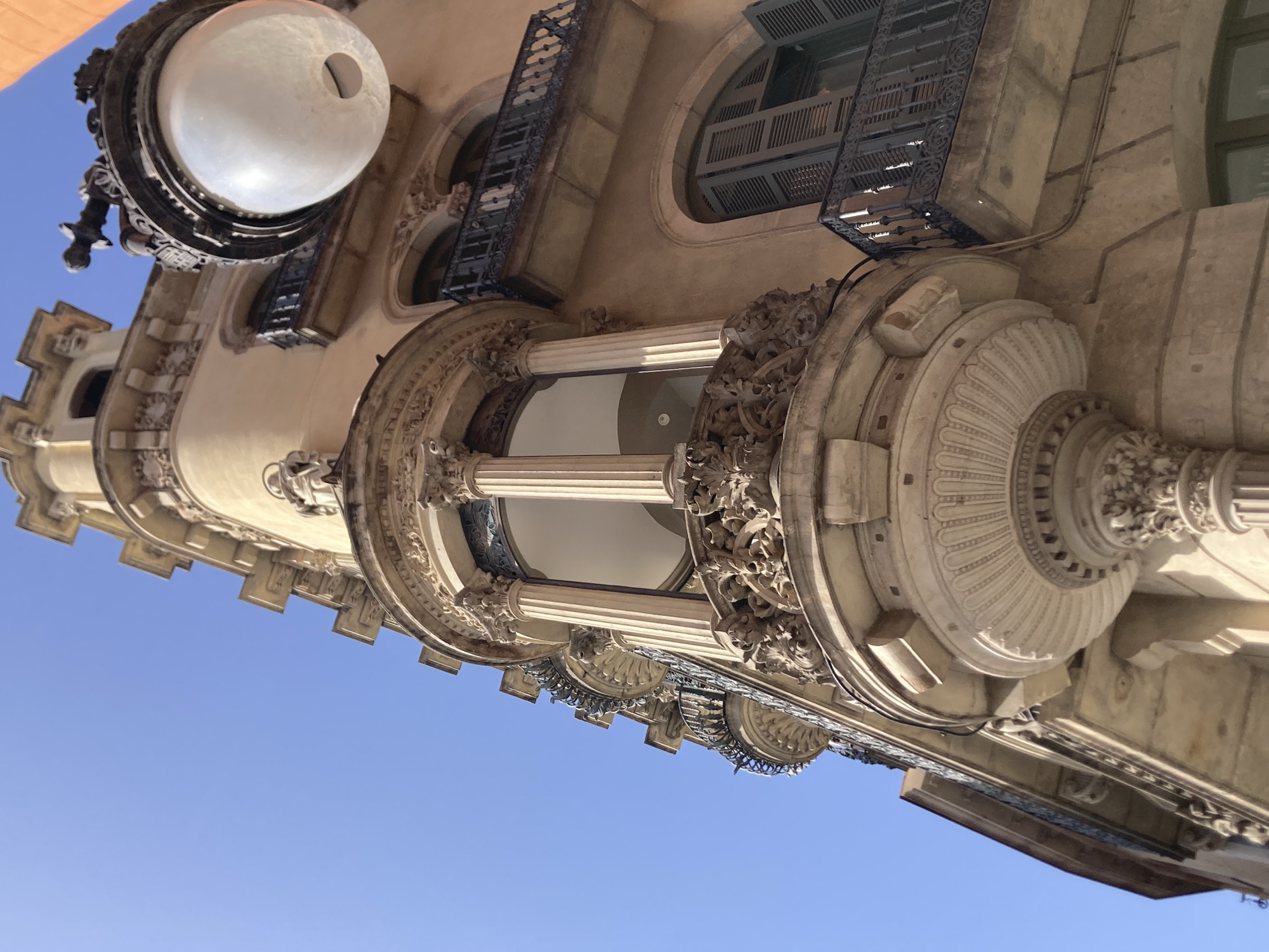 This is a low-angle, close-up, exterior shot of a building in Barcelona, Spain, featuring ornate architectural details. The building is made of a light-colored stone, possibly sandstone, and has a distinctly European style with decorative moldings, balconies with wrought-iron railings, and arched windows. A prominent feature is a rounded bay window with ornate carvings and columns, suggesting an Art Nouveau or Beaux-Arts influence, common in the Eixample district of Barcelona. Above the bay window, a small statue is visible. A decorative lamppost with a frosted glass globe is affixed to the exterior of the building in the upper right quadrant of the frame. The sky is a clear, bright blue, indicating a sunny day. No people are visible in the frame.