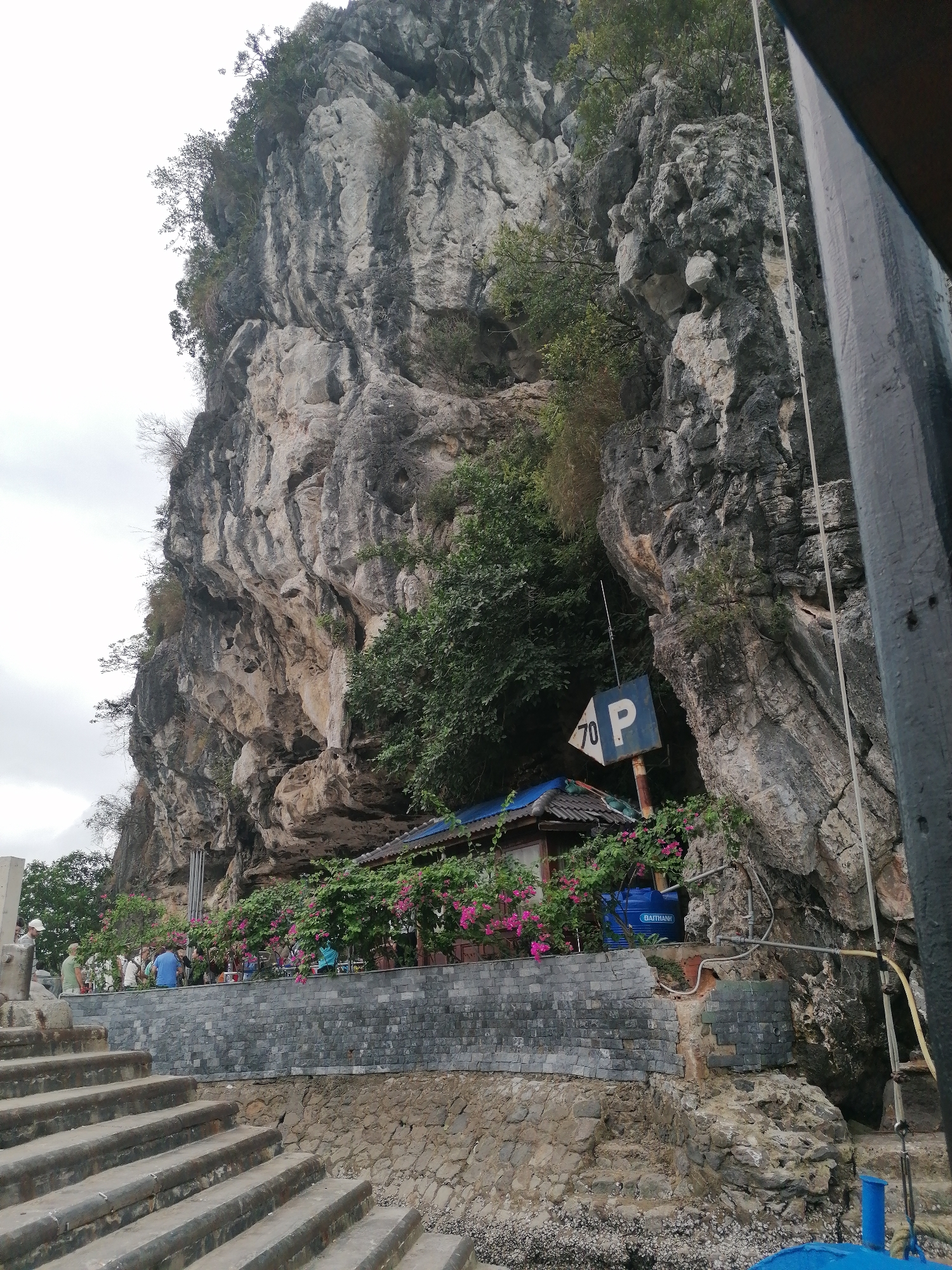 This media file presents a scenic view of a towering, rugged limestone karstic cliff, a geological feature typical of Ha Long Ward, Vietnam. The immense rock face, textured in shades of grey and tan, is partially covered with lush green vegetation, particularly at its summit and in various crevices.
At the base of the cliff, a small, rustic building with a blue corrugated metal roof is nestled amidst vibrant pink bougainvillea flowers. Adjacent to this structure, a blue water tank with the word "BAITHANH" visible on its side is positioned. A weathered blue and white sign, partially obscured but clearly displaying "70 P", is attached to the cliff face near the building.
Below the structure, a substantial grey brick retaining wall forms a terrace where several people are gathered. Individuals, dressed in casual clothing, are seen standing and sitting, appearing to be tourists or visitors observing their surroundings. Their expressions are not clearly discernible, but they seem relaxed.
In the immediate foreground, a series of wide, concrete steps descends towards a rocky, barnacle-encrusted shoreline or an embarkation point. The right side of the image is framed by dark wooden and metal elements, suggesting the vantage point is from a boat or a pier.
The sky is overcast, indicating a cloudy daytime setting. The scene suggests a popular natural attraction, possibly a stop for boat excursions or a viewing area, where human structures and activity are integrated with the dramatic natural landscape of the bay.