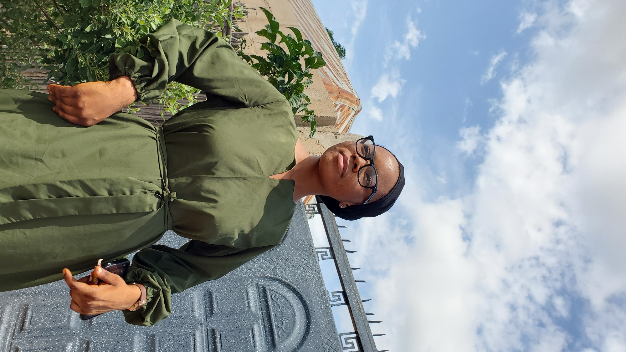 A young woman stands outdoors, facing right, with her hands on her hips. She is wearing a black headscarf, black-rimmed glasses, and a long, olive-green dress with puffed sleeves. The dress is cinched at the waist with a matching fabric belt. Behind her, a grey metal gate with decorative elements is visible. To her left, there are some green plants and a building with a corrugated metal roof. The sky above is blue with scattered white clouds. The scene appears to be in a residential area in Jalingo, Nigeria.
