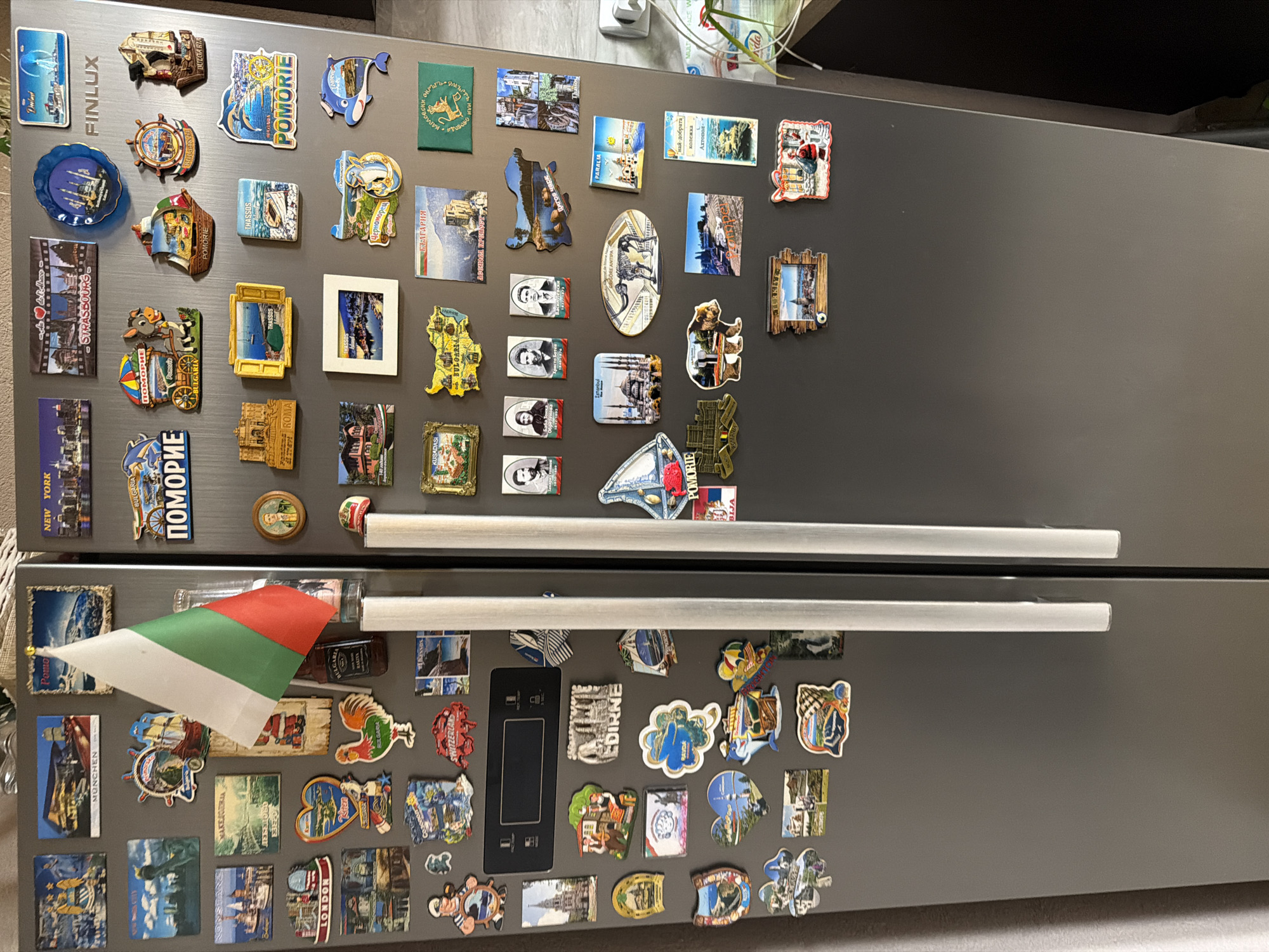 The image shows a gray refrigerator covered with souvenir magnets. The magnets feature various locations, including London, Munich, New York, Edirne, and various cities in Bulgaria such as Pomorie, Burgas, Karlovo and Panagyurishte (indicated by the magnet with "Карловски окръгъл" which references the region). There is also a small Bulgarian flag stuck to the refrigerator door.