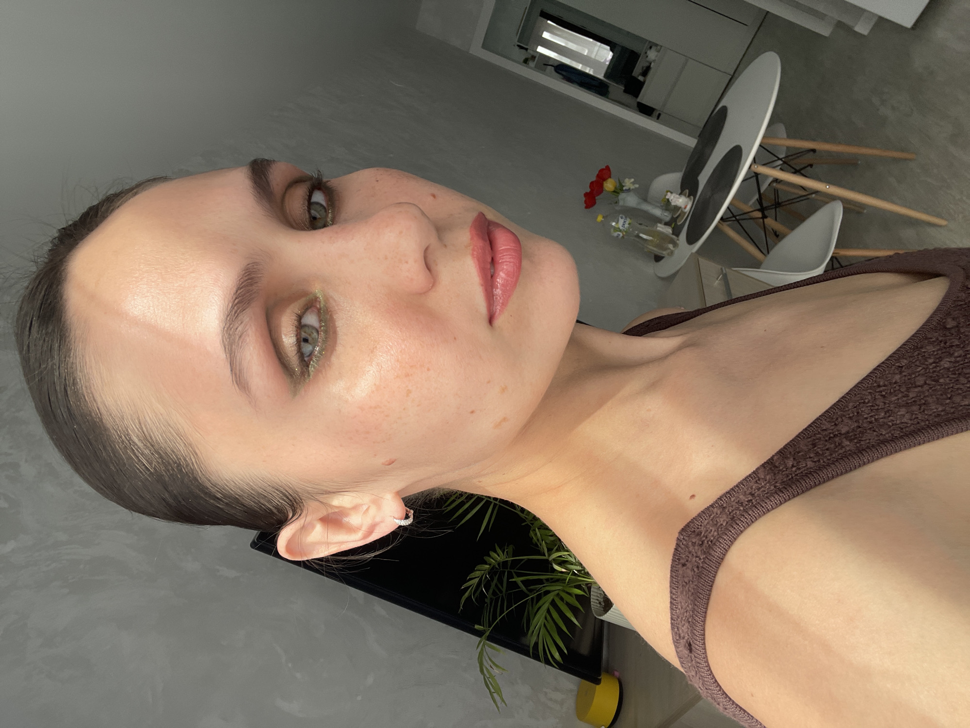 A medium shot depicts a fair-skinned woman with green eyes, slicked-back brown hair, and freckles. She is wearing light makeup, including green eyeshadow and pink lipstick, and has a small hoop earring in her left ear. She is wearing a brown ribbed top.
Behind her is an interior space with a gray wall to the left and a minimalist dining area. In the background, there is a white table with black accents, surrounded by white chairs with wooden legs. A vase containing red and white flowers is on the table, next to a bottle. Further back, a mirror reflects a bright room with a window. In the bottom of the frame, a plant with green leaves is visible, next to a yellow object. The overall lighting is bright and natural. The location is not explicitly identifiable as Izhevsk, Russia, based on the image alone.