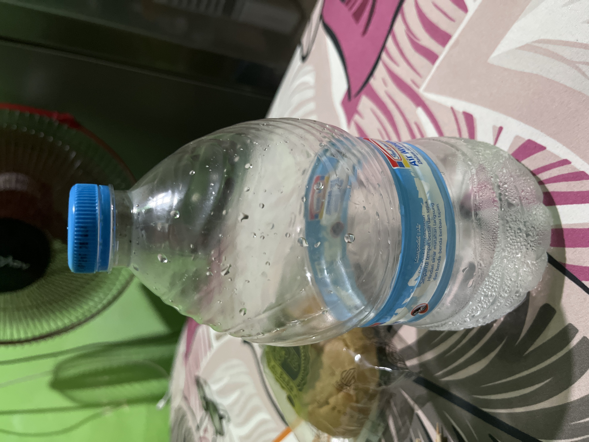 In Surabaya, Indonesia, a clear plastic bottle of drinking water sits on a patterned fabric surface. The bottle is partially filled with water, and condensation is visible on its exterior, suggesting a cool temperature. A blue plastic cap seals the bottle. To the left of the bottle, a round, green-wrapped item is partially visible, possibly a snack or food item. In the background, a green fan is partially visible, and the overall environment appears to be indoors. The lighting suggests the image was taken during the day or with artificial lighting.