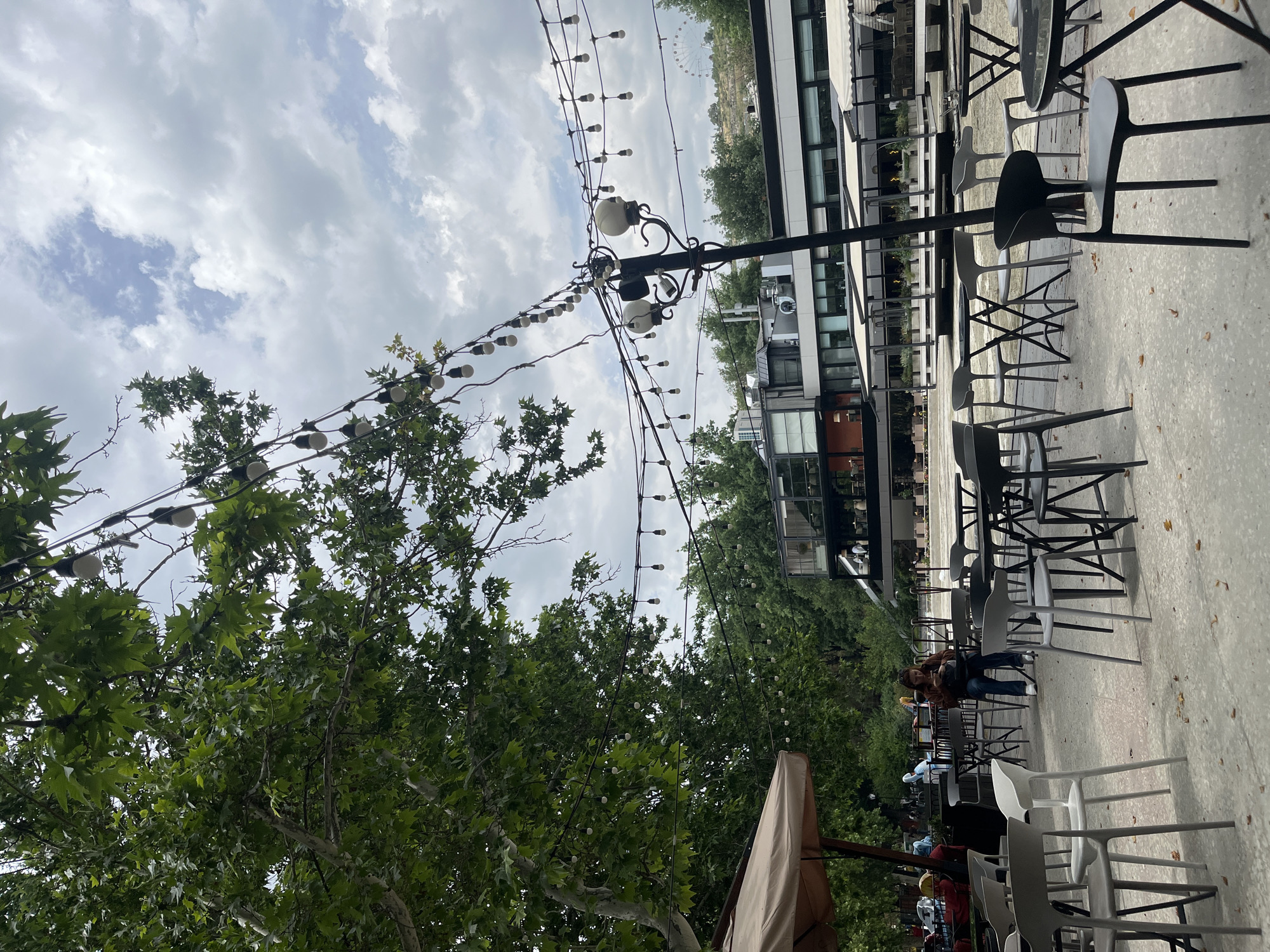 The image depicts an outdoor cafe scene in Yerevan, Armenia. Rows of empty black and white plastic chairs are arranged around tables on a paved surface, likely a patio or sidewalk. A few people are seated in the distance. Above, decorative string lights are strung between trees and lamp posts. A modern building with glass windows and metal accents is situated in the background. The sky is overcast with clouds. The image is rotated slightly.