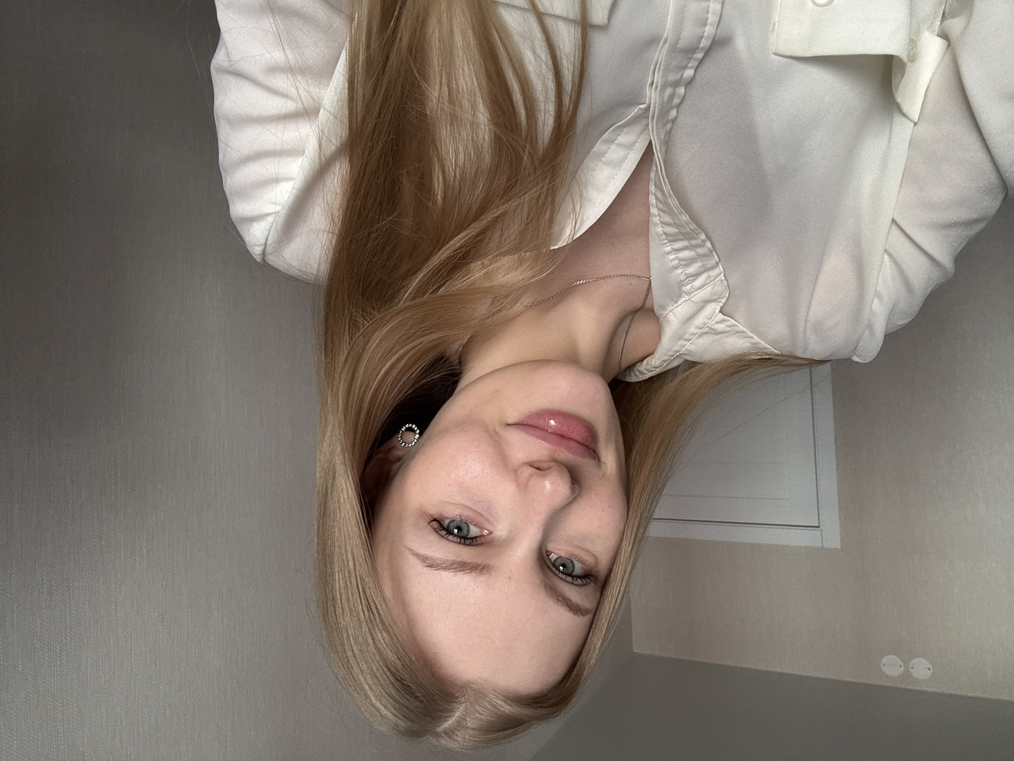 A young fair-skinned woman with long blonde hair is pictured indoors against a neutral-toned wall. She wears a white button-down shirt, partially unbuttoned, revealing a delicate silver necklace. Her eyes are light-colored, possibly blue or green. She has a small silver earring in her left ear. The lighting is soft and diffused. In the background, there is a white rectangular panel on the wall, along with two circular electrical outlets. There is no indication in the image that the photo was taken in Perm, Russia.