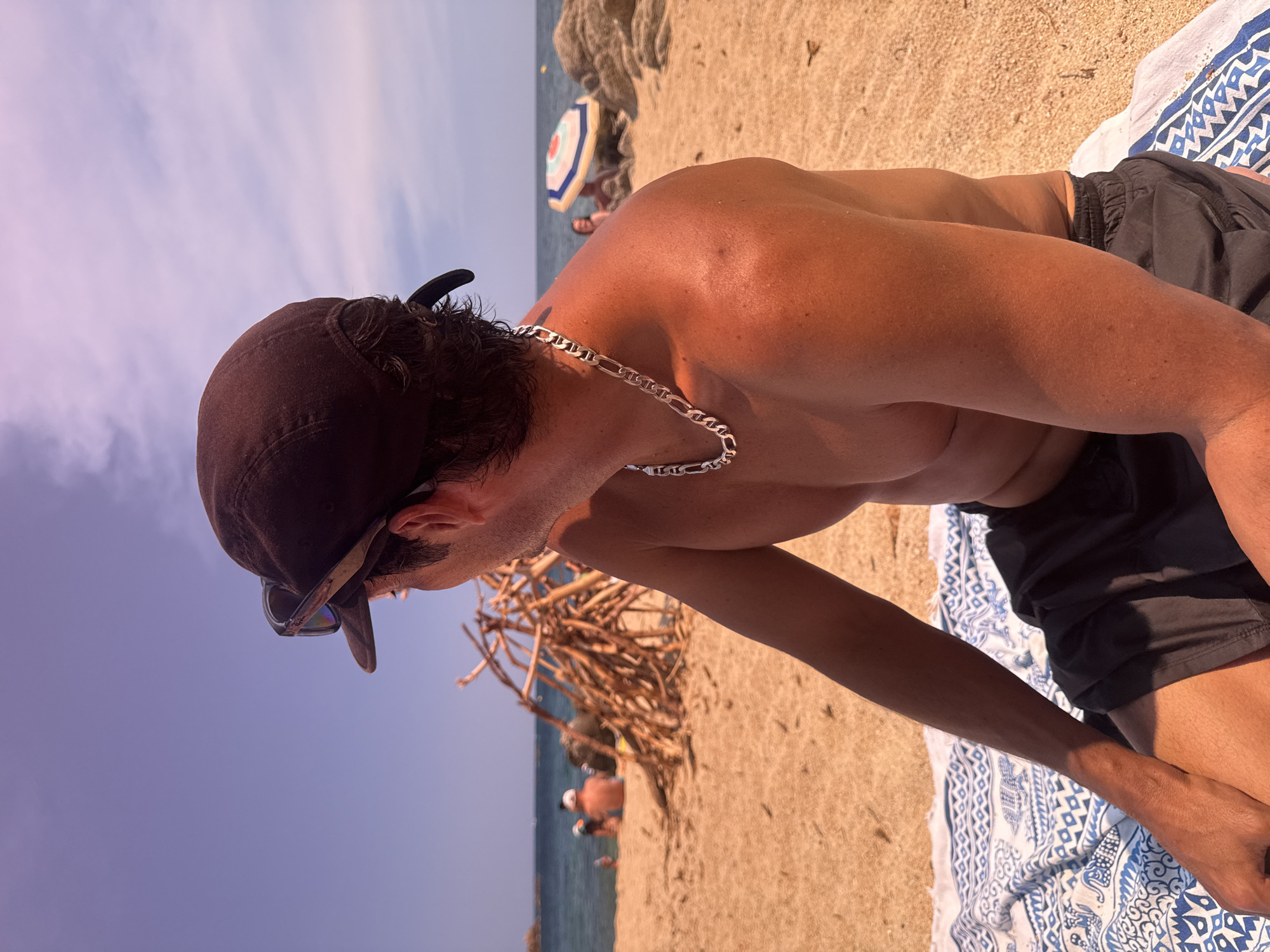 A man with a brown baseball cap and sunglasses perched on top is sitting on a beach in Calonge i Sant Antoni, Spain. He is facing away from the camera, looking towards the ocean. He is wearing a silver chain around his neck and dark shorts. He is sitting on a blue and white patterned towel on the sand. In the background, other people are visible on the beach, some sitting near a pile of driftwood and others near a colorful umbrella. The ocean is calm, and the sky is clear blue with some light clouds.