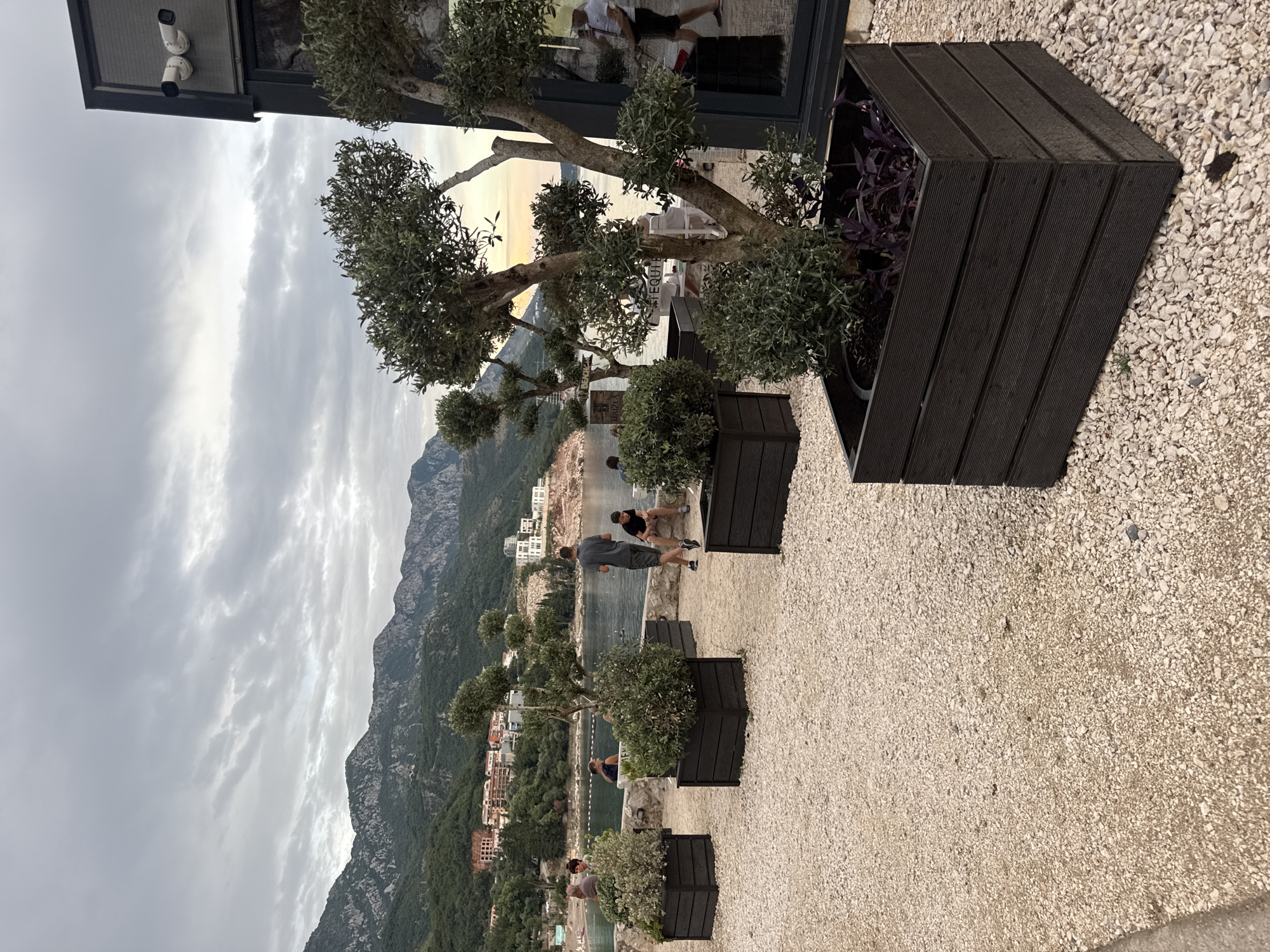 The image shows an outdoor scene in Kamenovo, Montenegro, likely a restaurant or cafe terrace overlooking the Adriatic Sea. The perspective is from a raised vantage point, looking down towards the water and a distant mountainous coastline. The foreground features a gravel path and several dark wooden planters containing greenery, including an olive tree.
Several people are visible in the mid-ground, appearing to be patrons enjoying the outdoor space. One couple sits on a low wall near the water, while a man in a grey shirt and shorts stands and looks towards the sea. Further down, another person is visible near a planter. The overall atmosphere seems relaxed and leisurely.
In the background, a body of water is visible, with boats docked along the shore. Beyond that, a town with white buildings nestles at the foot of lush, green mountains that rise steeply from the coast. The sky is overcast, with patches of brighter sky suggesting either early morning or late afternoon light. The weather appears to be mild and possibly a bit cloudy.
A few details stand out: two white security cameras are mounted on an overhang in the upper left corner of the image. Inside a windowed structure to the right of the cameras, a reflection shows people and furniture, suggesting the presence of an indoor seating area.
No discernible text is present in the image. The scene conveys a sense of calm and natural beauty, with the built environment integrated into the picturesque landscape.
