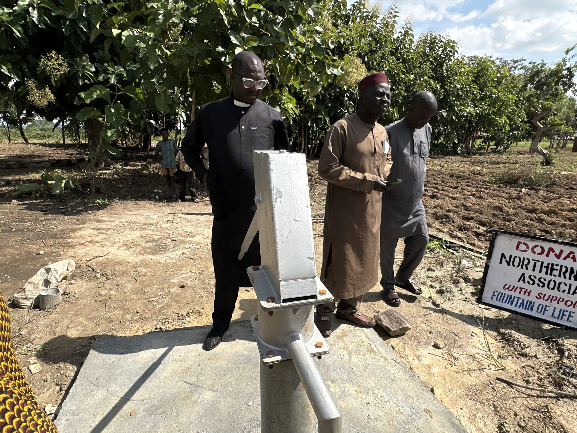 In Afogo, Nigeria, three men stand near a water pump. The man on the left, wearing glasses and a black robe with a white collar, stands closest to the pump. To his right, a man in a brown robe and a red hat is holding a document. The third man, in a grey robe, stands slightly behind and to the right of the man in brown. The water pump is a metallic structure mounted on a concrete base. In the background, there are trees and tilled earth. A sign with red and blue lettering is visible on the right side of the image. The ground is dry and dusty.