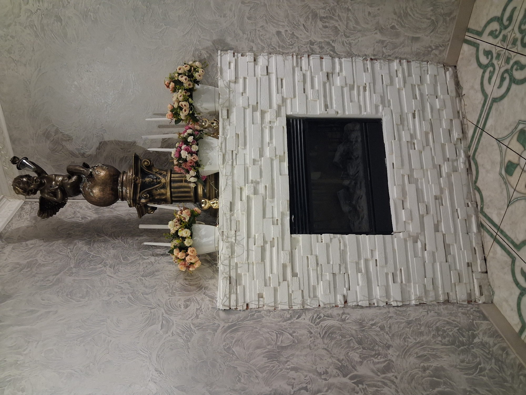 The image shows an interior scene featuring a decorative fireplace. The fireplace surround is constructed from white, irregularly shaped and sized rectangular blocks, giving it a textured, stacked stone appearance. A black insert denotes the fireplace opening. Above the fireplace, on the mantelpiece, are two white pots filled with clusters of light pink and white flowers. Behind the floral arrangements, three white tapered candles stand upright. A bronze statue of a cherub is positioned on a decorative column to the left of the fireplace, holding a torch. The wall behind the fireplace is decorated with a textured gray wallpaper that features a swirling, abstract pattern. The floor is tiled with a pattern of light and dark green, ornate designs on a cream background. The scene is lit, highlighting the decorative elements. The location is identified as being in the city of Znamianka, Ukraine.