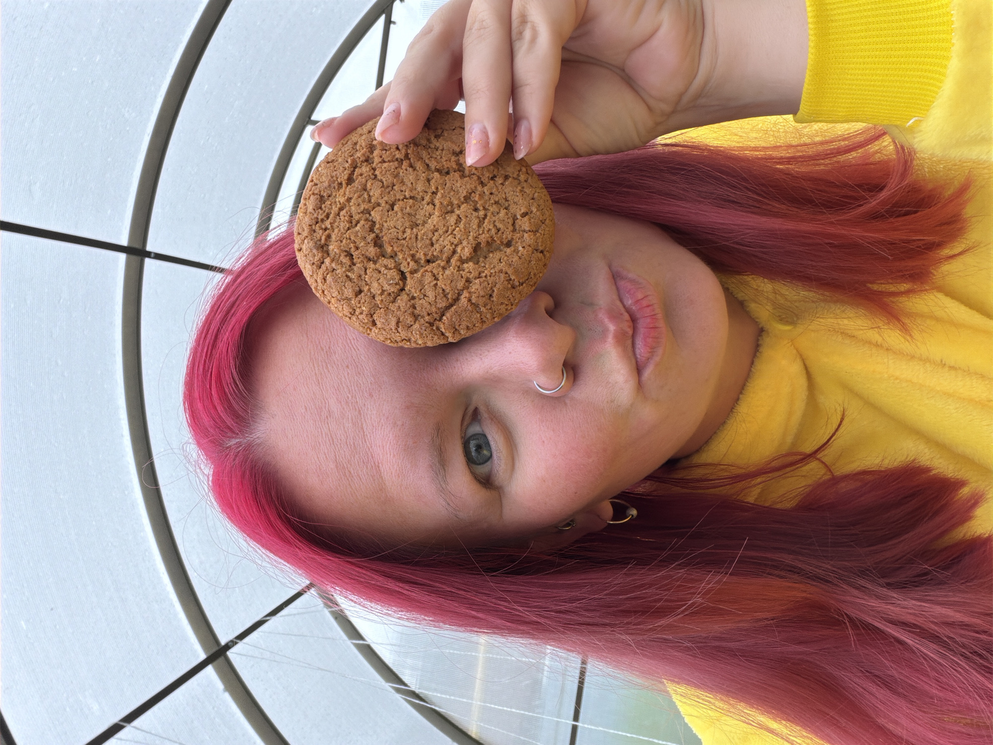 An individual with bright fuchsia-colored hair and fair skin is depicted in a close-up portrait within a translucent, arch-framed structure, resembling a greenhouse, located in Chaikovsky, Russia. The individual's visible left eye is blue. A round, light-brown cookie is held by the individual's right hand, obstructing the right side of the face. The individual wears a silver hoop nose ring in the left nostril and small hoop earrings in both ears. The lips are positioned in a puckered gesture. The individual is dressed in a textured, bright yellow long-sleeved garment, possibly fleece. The background consists of diffused natural light filtered through a translucent ceiling, revealing dark grey structural arches and intersecting lines, along with thin white wires. The fingernails of the hand holding the cookie are covered with a light, semi-transparent polish featuring small, subtle flecks.