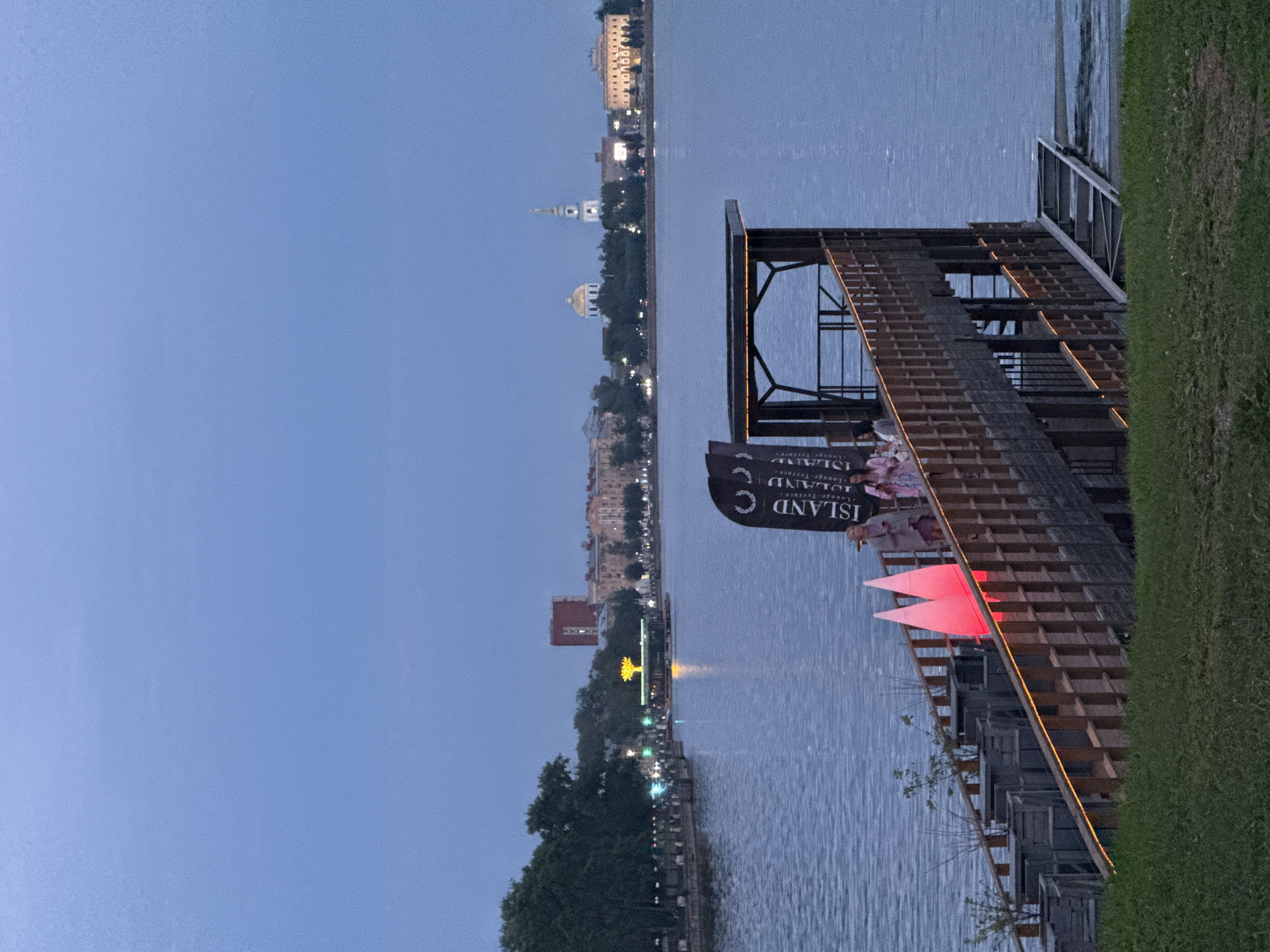 The image depicts a twilight scene at the embankment of Votkinsk, Russia. A wooden pier or boardwalk extends into a body of water, illuminated by warm orange lights along its railings. On the pier, several people are visible, some standing and others appearing to walk. Two triangular, pinkish-red illuminated structures are also present on the pier. In the background, across the water, the Votkinsk skyline is visible, featuring buildings with lights, including what appears to be a church with a steeple and a domed building. Trees line the embankment in the mid-ground, and a floating structure with a decorative tree-like light installation is in the water. The overall atmosphere suggests an evening event or gathering.