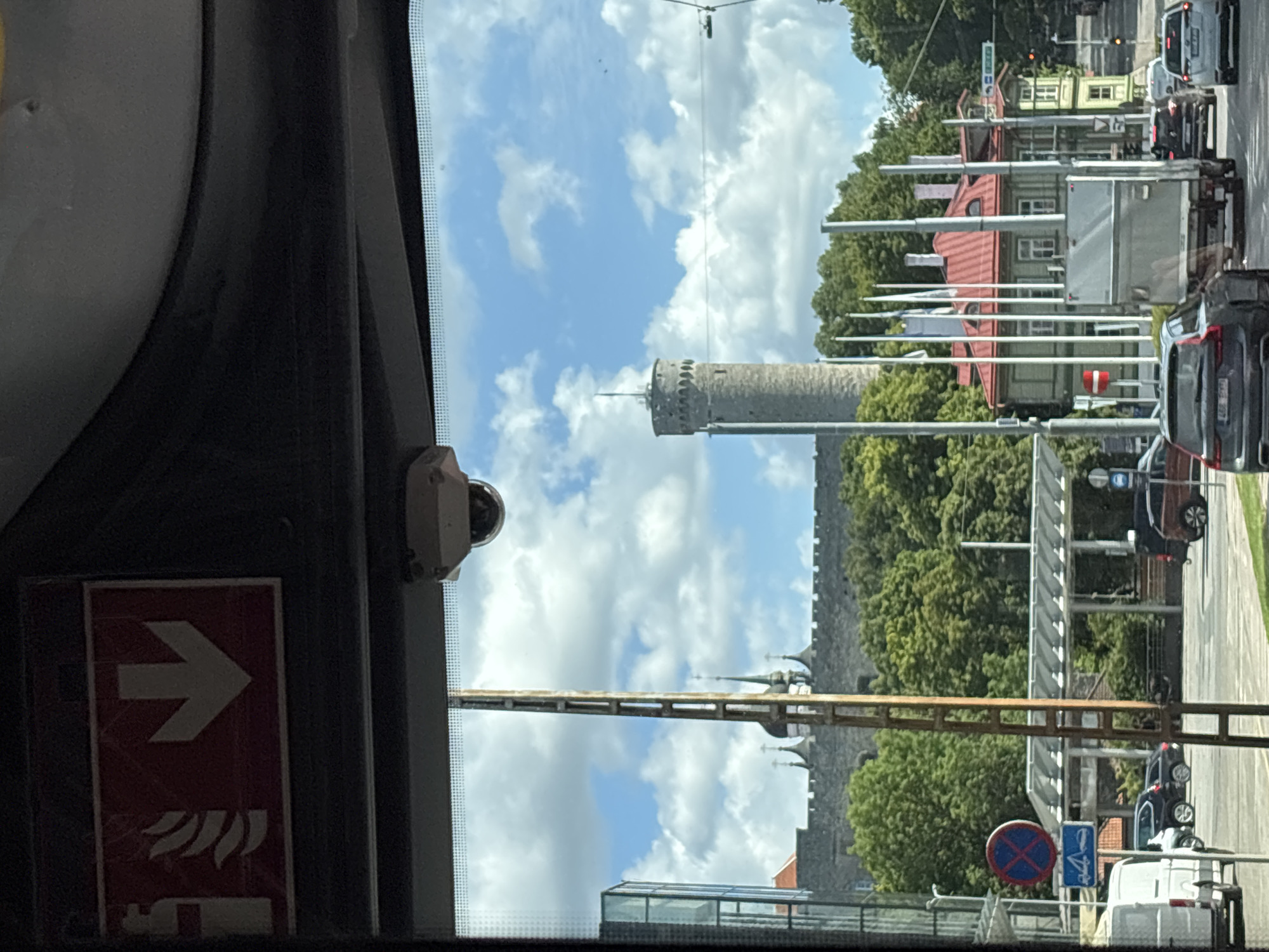 The image is taken from inside a vehicle looking out through the windshield. At the top of the frame, a red and white fire safety sign with a downward pointing arrow is visible. Below this, a small security camera is mounted. The foreground shows a street scene with vehicles, including a white van on the left and several cars. A large, rough-hewn stone wall with battlements stretches across the mid-ground, with several church spires peeking over it. To the right of the wall, a tall, cylindrical stone tower with a crenellated top and a spire stands prominently against a blue sky with scattered white clouds. In the foreground, several flags on tall poles are planted in front of a red-roofed wooden building. The scene appears to be in Tallinn, Estonia, likely depicting the historic Old Town area with its defensive walls and iconic towers.