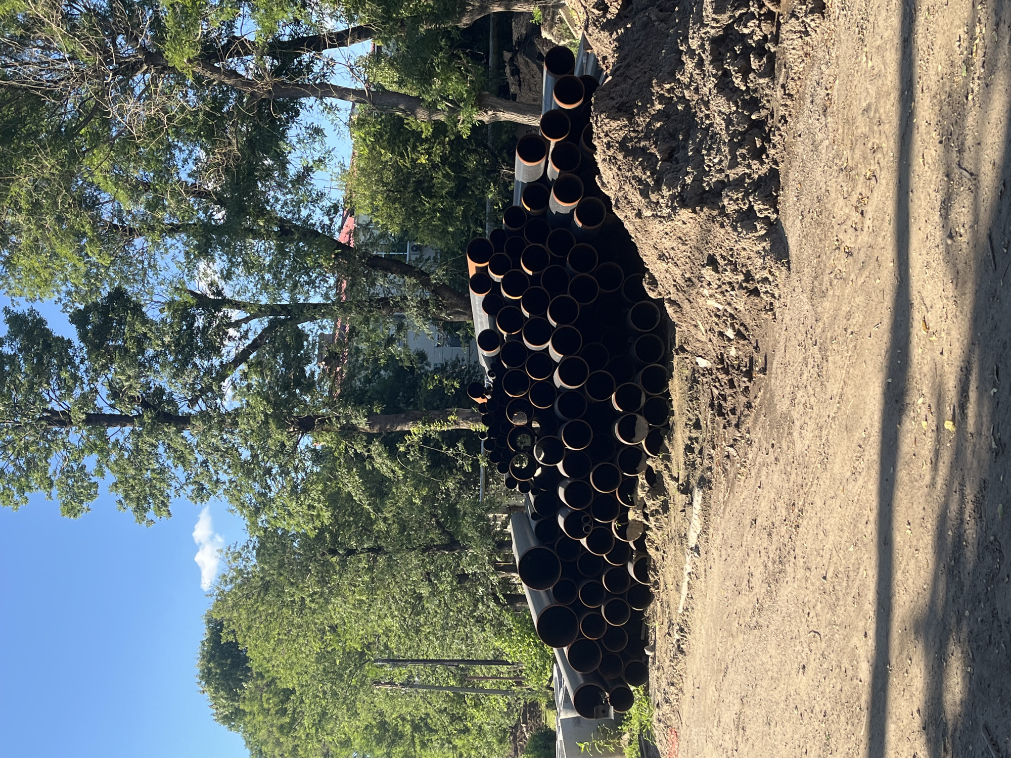 A pile of large black pipes lies on a dirt surface in front of a line of trees and a building. The pipes are cylindrical with open ends, and they are stacked haphazardly, with some lying horizontally and others at an angle. The dirt surface is uneven and appears to be a construction site. The trees are lush and green, and the sky is a clear blue with a few scattered clouds. The building in the background is light-colored with a red roof. This scene is consistent with an outdoor construction site in Krasnodar, Russia.
