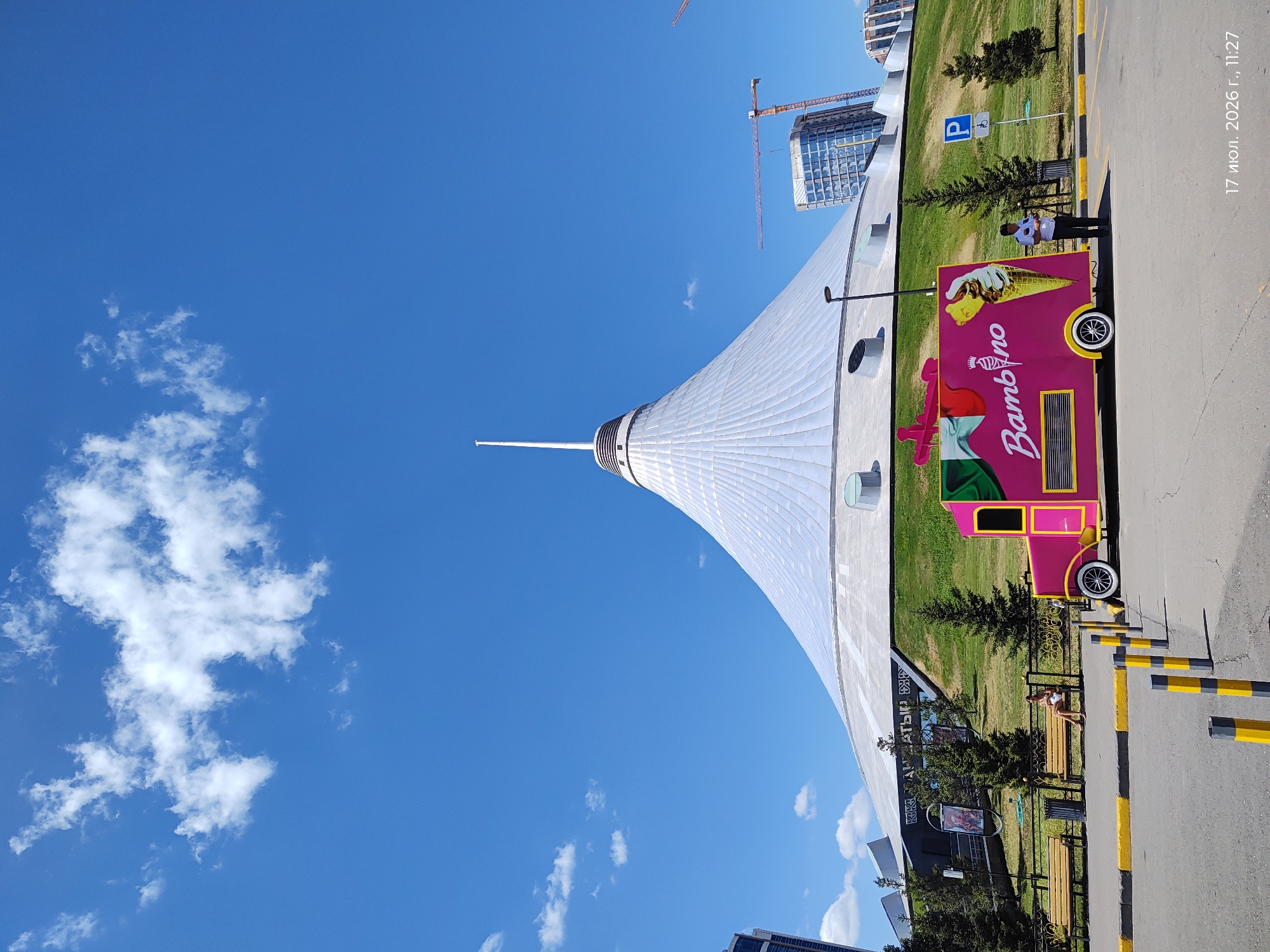 The image depicts a bright, sunny day in Astana, Kazakhstan, with a clear blue sky and scattered white clouds. Dominating the background is the Khan Shatyr Entertainment Center, a distinctive tent-shaped building with a tall spire. Its metallic exterior reflects the sunlight. In front of the building, a grassy incline leads down to a paved area.
In the foreground, a vibrant pink ice cream truck, branded with "Bambino" and featuring illustrations of ice cream, is parked. To the left of the truck, two wooden benches are visible, with one occupied by a woman. A trash receptacle is next to the bench. A black and yellow striped bollard-lined pathway separates the paved area from the grassy slope. A parking sign with a wheelchair symbol is visible on the right side of the image. A person, possibly an employee, stands near the ice cream truck. In the distance, construction cranes and modern buildings are visible, indicating ongoing development in the city. A timestamp in the bottom right corner reads "17 июл. 2026 г., 11:27".