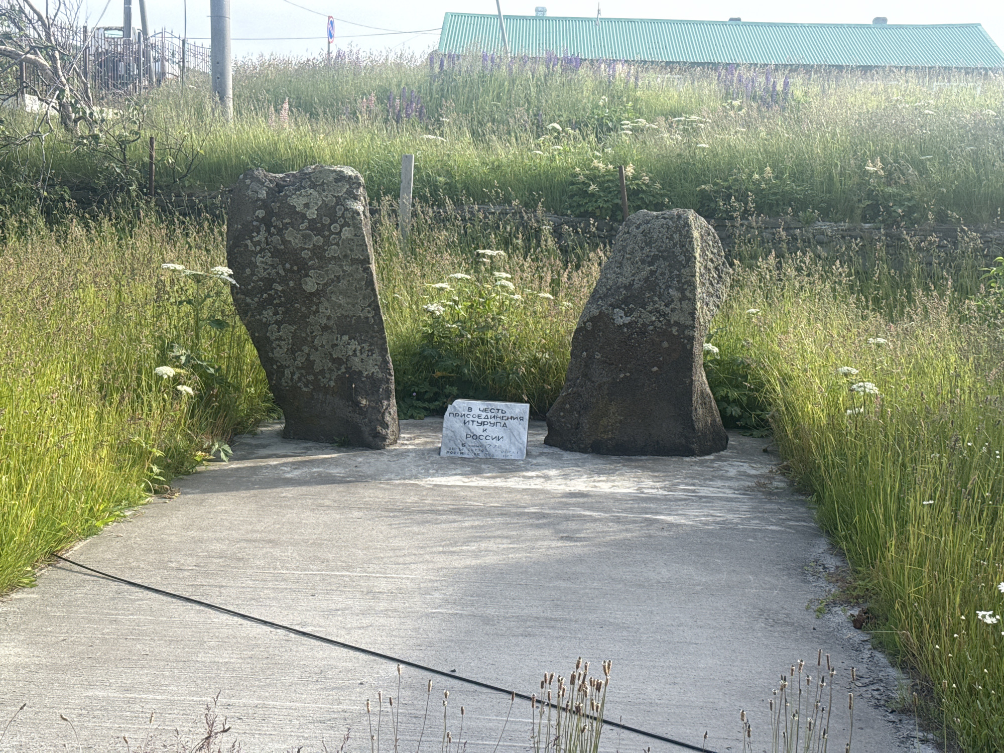 The image depicts a memorial consisting of two large, weathered stones standing upright on either side of a concrete pathway. Between the stones, a white stone plaque is placed on the ground, bearing Cyrillic inscription. The plaque reads "В ЧЕСТЬ ПРИСОЕДИНЕНИЯ ИТУРУПА РОССИИ" ("In honor of the incorporation of Iturup to Russia"), with smaller text below indicating dates and possibly names. The pathway is bordered by tall, green grass and wildflowers, with a dense field of vegetation and a distant building with a green corrugated roof in the background. A dark wire runs diagonally across the concrete in the foreground. The scene appears to be an outdoor memorial site in a natural setting, possibly in or near Kuril'sk, Russia, given the inscription on the plaque.