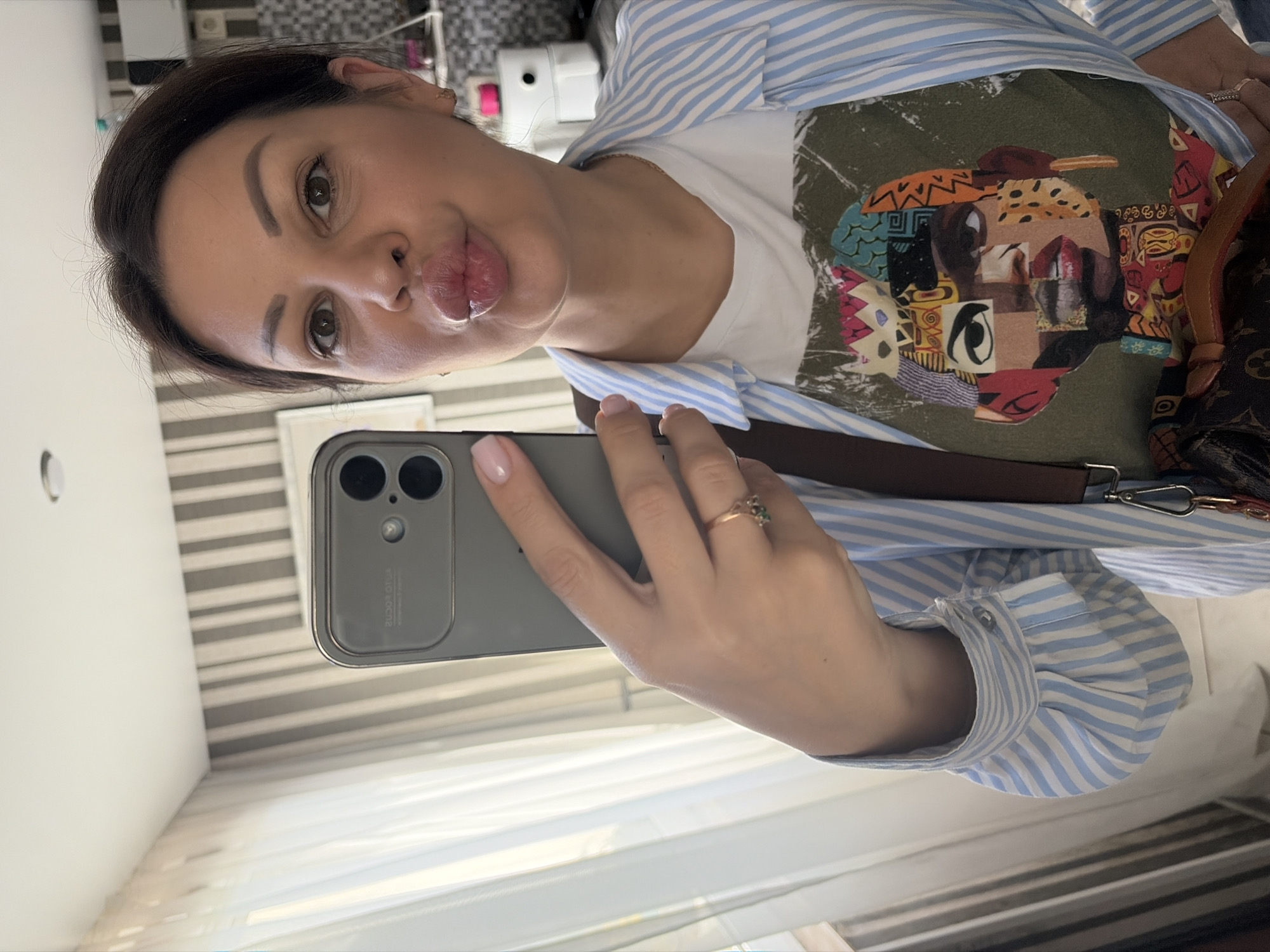 The image displays a woman taking a selfie indoors in Sochi, Russia. The woman, with dark hair pulled back, brown eyes, and light makeup, faces the camera with puckered lips. She holds a dark gray smartphone with a dual-camera setup in her right hand; her fingernails are painted light pink, and a gold-toned ring with green stones is on her right ring finger. She wears a white t-shirt featuring a multi-colored, abstract graphic print of a human face, layered under an unbuttoned light blue and white striped long-sleeved shirt. A brown strap from a Louis Vuitton monogram bag is visible over her right shoulder, with a portion of the bag at the bottom right. The background on the left shows sheer white curtains over a window, while the right side features a wall with light gray and white vertical stripes, transitioning to a tiled area with a white appliance and a pink-capped object. The word "AUTO FOCUS" is visibly reversed on the phone's camera module, indicating a reflection.