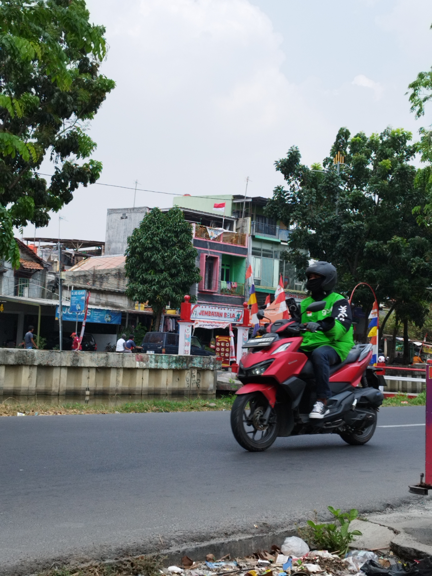 A person wearing a green jacket and a black helmet rides a red motorcycle along a paved road. The motorcycle has a black seat and black wheels. The rider's legs are visible, clad in blue jeans, and they are wearing white and black shoes. The background shows buildings of varying heights and colors, some with balconies and flags. Trees are present, some with dense green foliage. A concrete barrier runs alongside the road, with water and vegetation visible beyond it. The road appears to be in an urban or suburban setting in Bekasi, Indonesia, as indicated by the signage and architectural style. The scene is captured during the day, with natural light illuminating the environment. The road surface shows some signs of wear and tear. Trash is visible along the edge of the road.