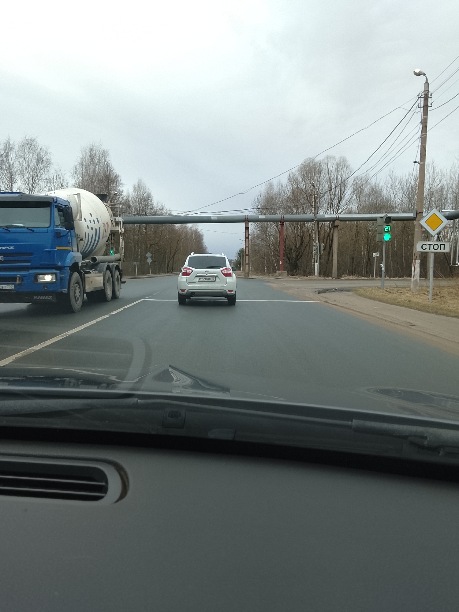 Captured from the interior of a car, the image shows a view of a road in the city of Protvino, Russia. The road is wet, suggesting recent rainfall. A white SUV with the license plate P228HB49 is directly ahead, and a blue KAMAZ cement mixer truck is on the left side of the road. Overhead, a set of pipes spans the road. To the right of the road, there is a traffic light displaying a green light and a "СТОП" (STOP) sign. Trees are visible in the background. The weather appears overcast. The dashboard of the car the image was taken from is visible in the foreground.