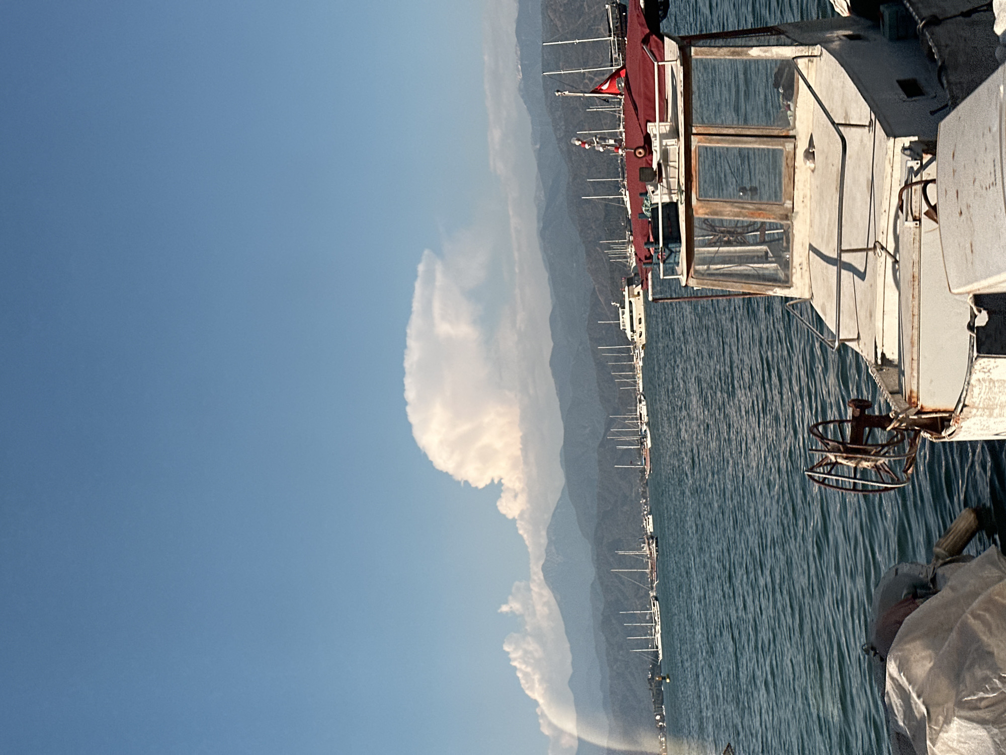 A panoramic view captures a sunny day along a mountainous coastline, likely in Fethiye, Turkey, given the presence of a Turkish flag. The image is taken from a boat, with parts of its structure framing the right side of the frame. In the immediate foreground on the right, a white boat cabin with wooden-framed windows is visible, along with a rusted steering wheel or winch, and a part of a red canvas covering the deck. A white tarp or cover is also seen at the very bottom right corner.
Beyond the boat, the calm blue sea stretches across the midground, showing gentle ripples on its surface. Along the distant shoreline, numerous other boats, possibly fishing vessels or yachts, are moored or anchored in a long line, with their masts and superstructures visible.
In the background, a series of majestic mountains rise, their peaks shrouded in a light, atmospheric haze. Above the mountains, the sky is a clear, vibrant blue, punctuated by a few bright white cumulus clouds. One prominent, fluffy cloud formation stands out against the blue sky on the left side of the image. The overall impression is one of a peaceful maritime scene under bright daylight. No discernible text is visible in the image.
