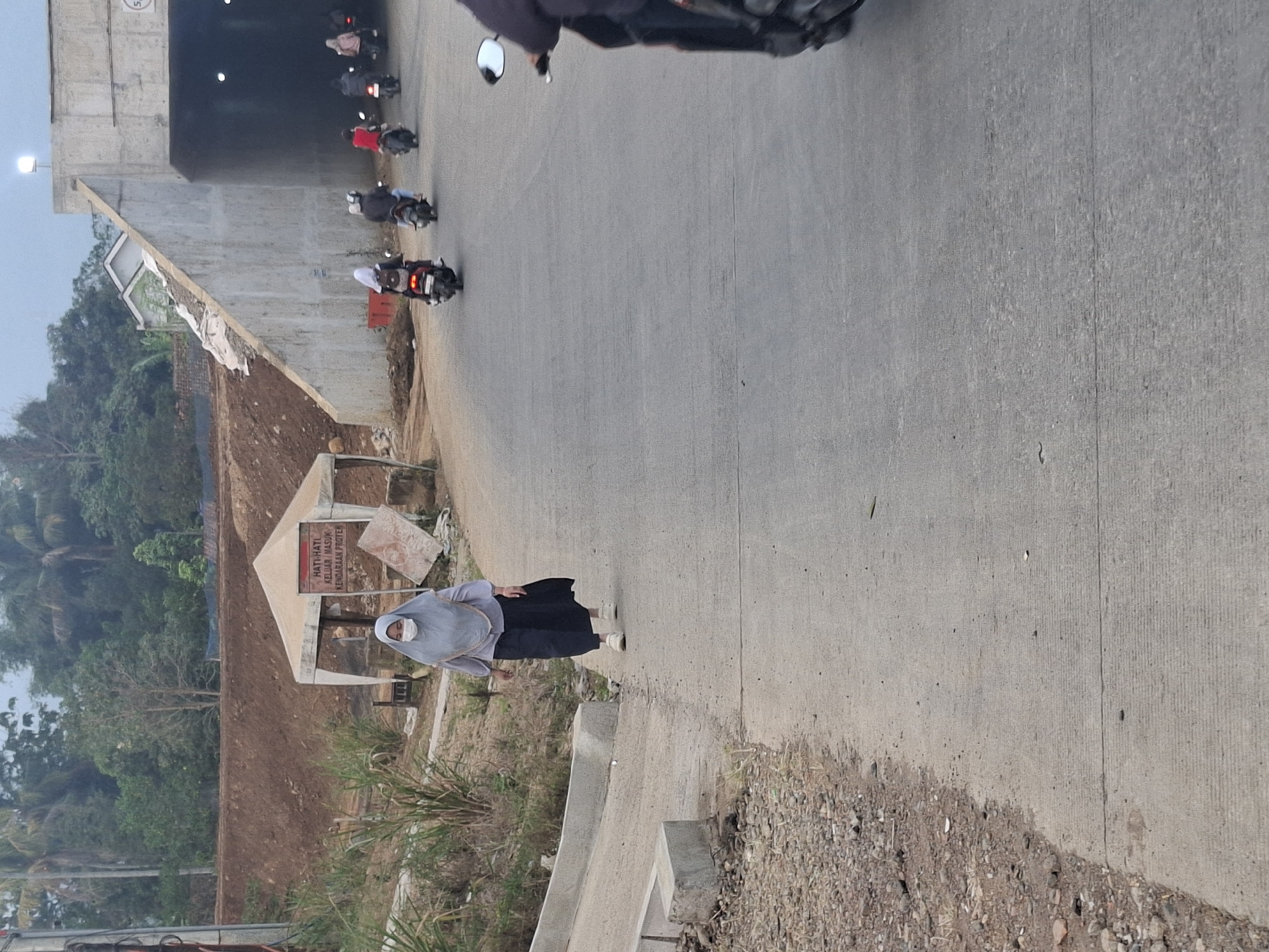 A woman wearing a grey hijab and face mask walks on a paved road near a construction site. She is wearing a grey long-sleeved shirt and a dark skirt, with white sneakers. In the background, there is a construction tent with a sign. To the right, a concrete tunnel entrance is visible, with several motorcycles and riders entering it. The environment appears to be a semi-urban or rural area with trees and earth mounds, suggesting ongoing construction or road development. The lighting suggests it is either early morning or late afternoon.