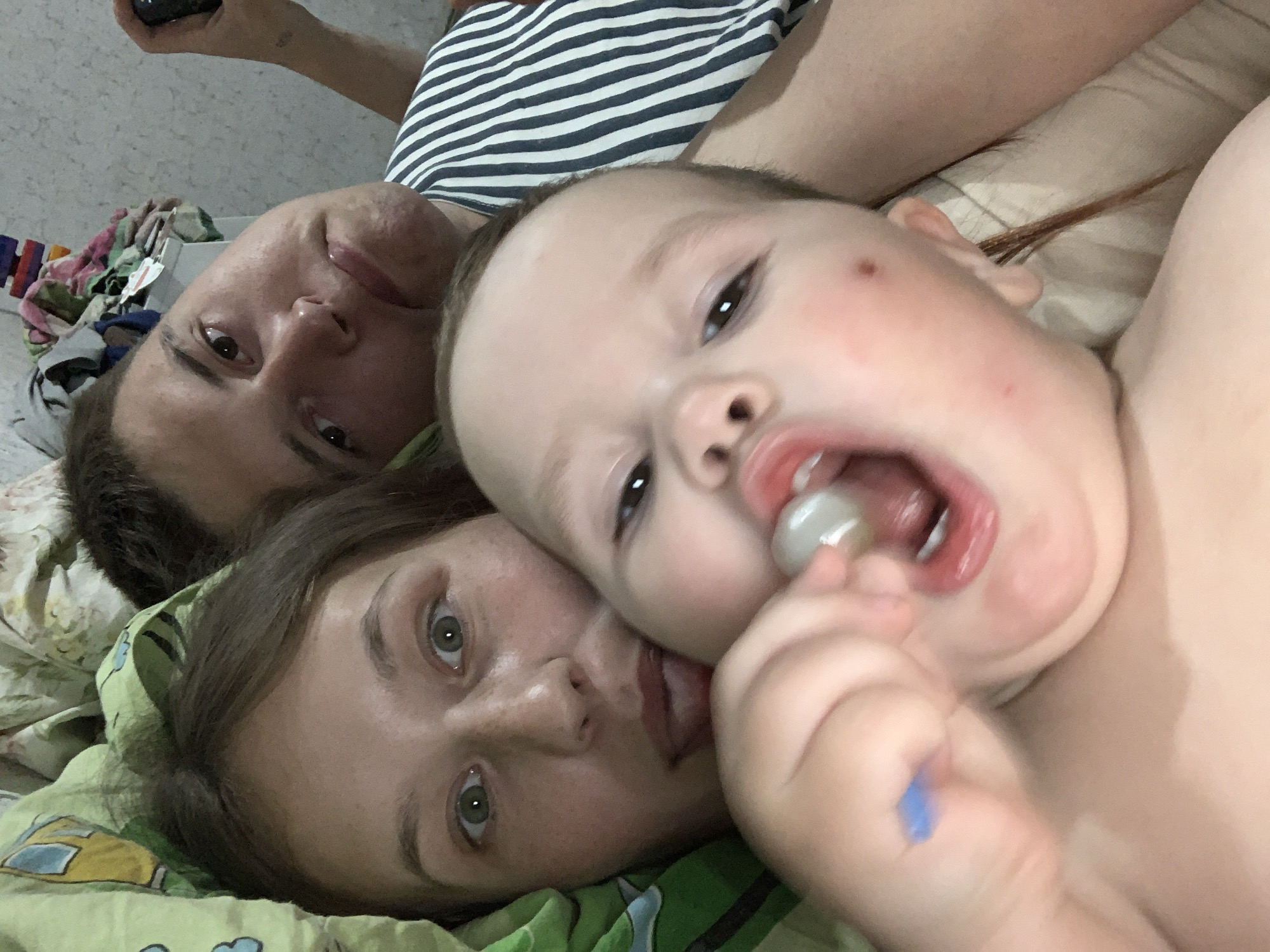 The image appears to be a selfie taken indoors, possibly in a private residence in Фоки, Russia. It captures three individuals: a man, a woman, and a baby. The man and woman are lying down, likely on a bed, with their heads close together. The baby is positioned closer to the camera, with an open mouth revealing a lollipop or similar treat. The man is wearing a striped shirt. The environment includes bedding with a green patterned sheet. The overall scene suggests a casual, intimate moment between family members.