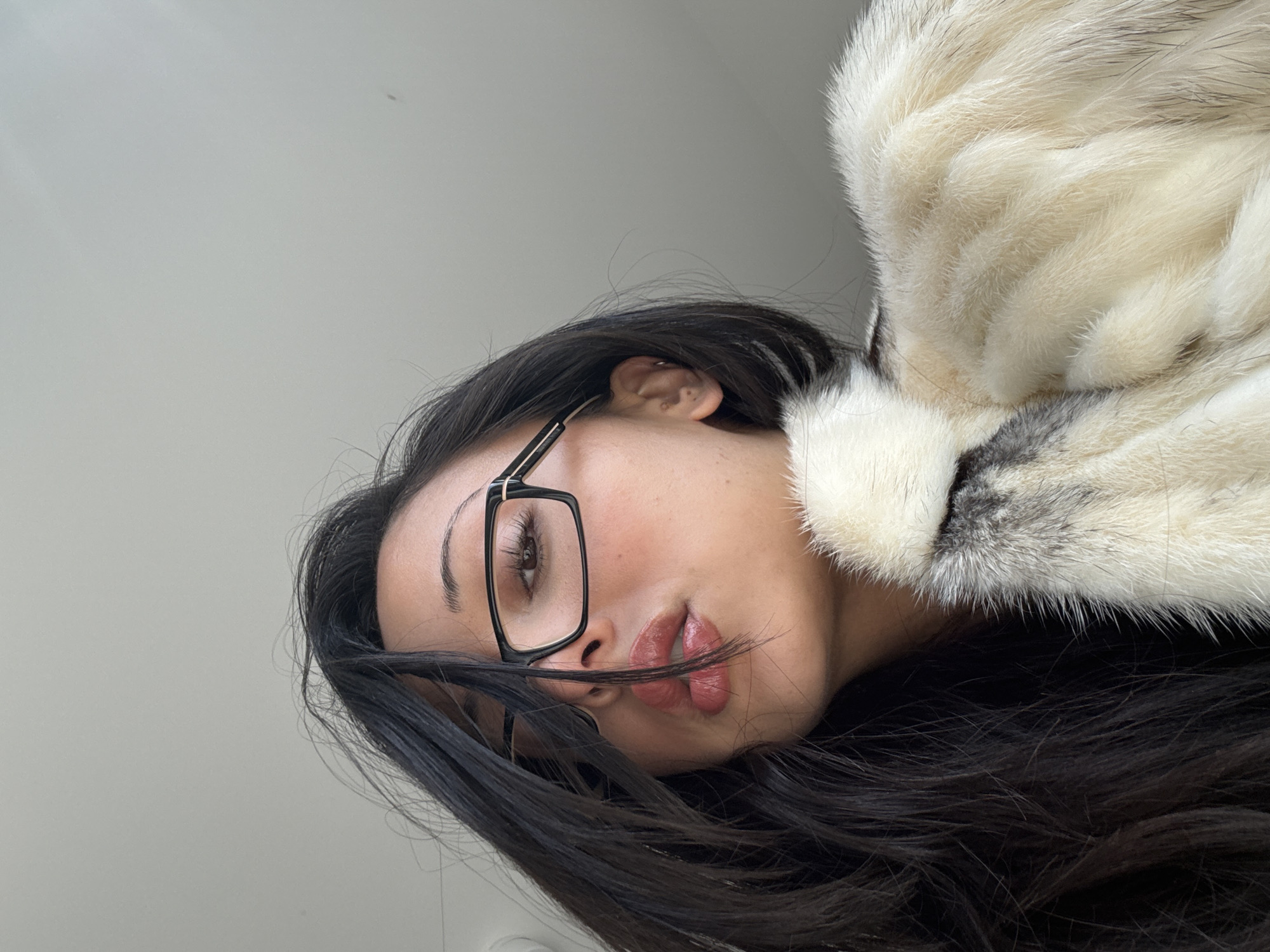 A portrait-style image, taken from a low angle, features a young woman posing indoors against a plain, light grey background. Her head is tilted to the right, suggesting she is lying down or leaning back. She has long, dark, flowing hair and is wearing prominent black-framed, rectangular glasses. Her eyes are open, looking towards the camera, with visible eyeliner. Her lips are full and slightly parted, with a hint of lip color, and a strand of her dark hair rests across her lower lip.
She is dressed in a fluffy, multi-toned garment, likely fur or faux fur, which appears white, grey, and black, adding a strong textural element to the image. A close-up view reveals the intricate texture of this garment and the fine details of her ear with a small piercing. The image captures a serene or contemplative moment, focusing on her facial features and the textural contrast between her hair and the garment. There is no visible text. The setting is an indoor environment in London, United Kingdom.