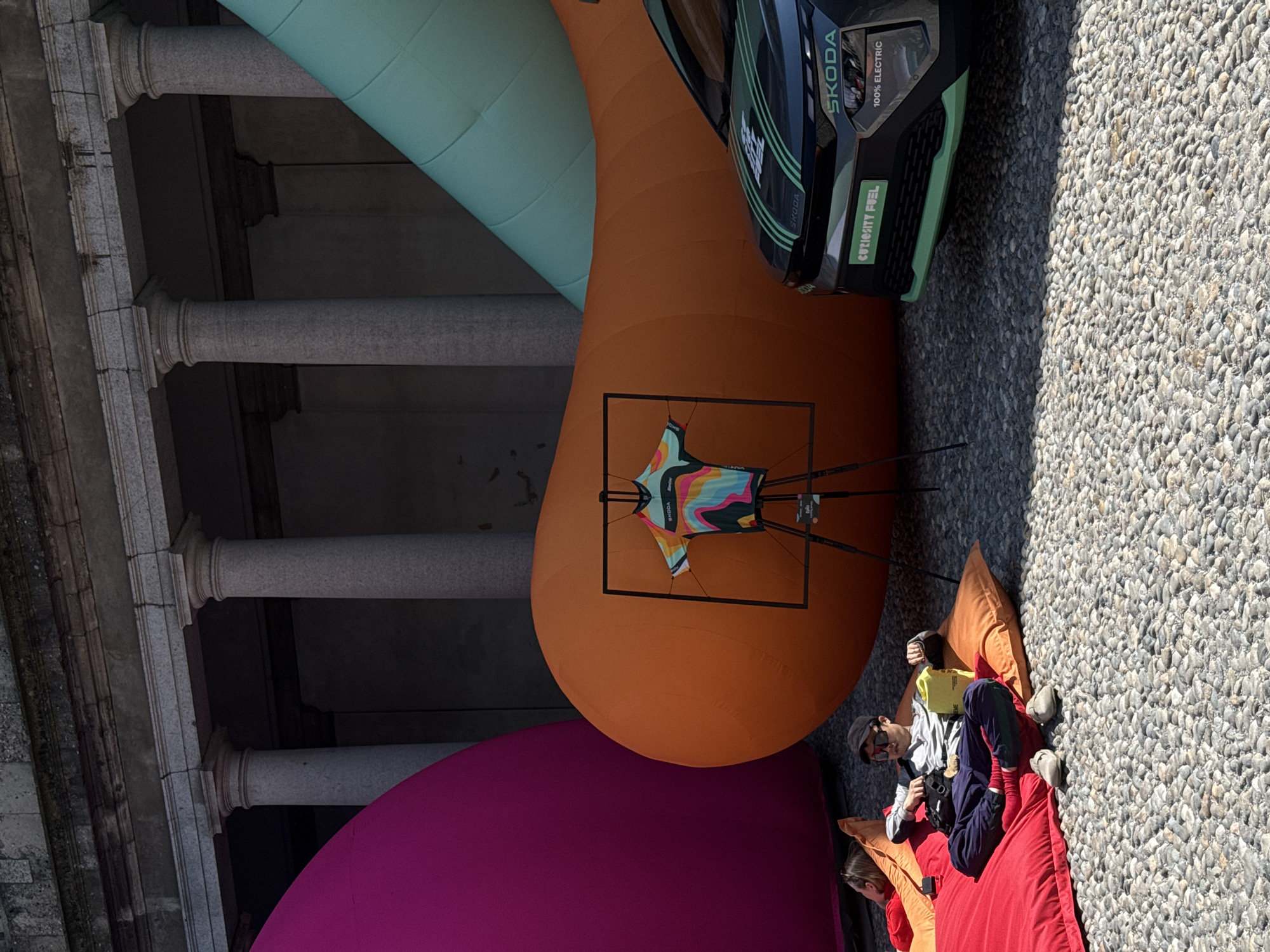 An outdoor scene in Milan, Italy, captures a vibrant installation or promotional event within a historic courtyard setting. Large, interconnected inflatable structures in bright colors dominate the space: a teal one extends from the upper left, a prominent orange one curves through the center, and a magenta one rests at the bottom left.
A dark grey/black Skoda electric car is positioned on the orange inflatable, with visible text reading "SKODA," "100% ELECTRIC," and "CURIOUSLY FUEL" on its front. Nearby, a colorful cycling jersey with an abstract pattern, displaying "SKODA" and partially "Racing," hangs on a black frame, also on the orange inflatable.
On the cobblestone ground to the right, two individuals are seen relaxing on large beanbag chairs, one orange and one red. A man wearing a cap and sunglasses sits on the red beanbag, holding a yellow bag. The head of another person is visible towards the bottom edge.
In the background, sturdy grey stone columns and a textured stone wall of an old building are prominent, contrasting with the modern installation. The scene is illuminated by bright daylight, suggesting sunny weather. The setting combines contemporary design with traditional architecture, creating a relaxed and engaging atmosphere.