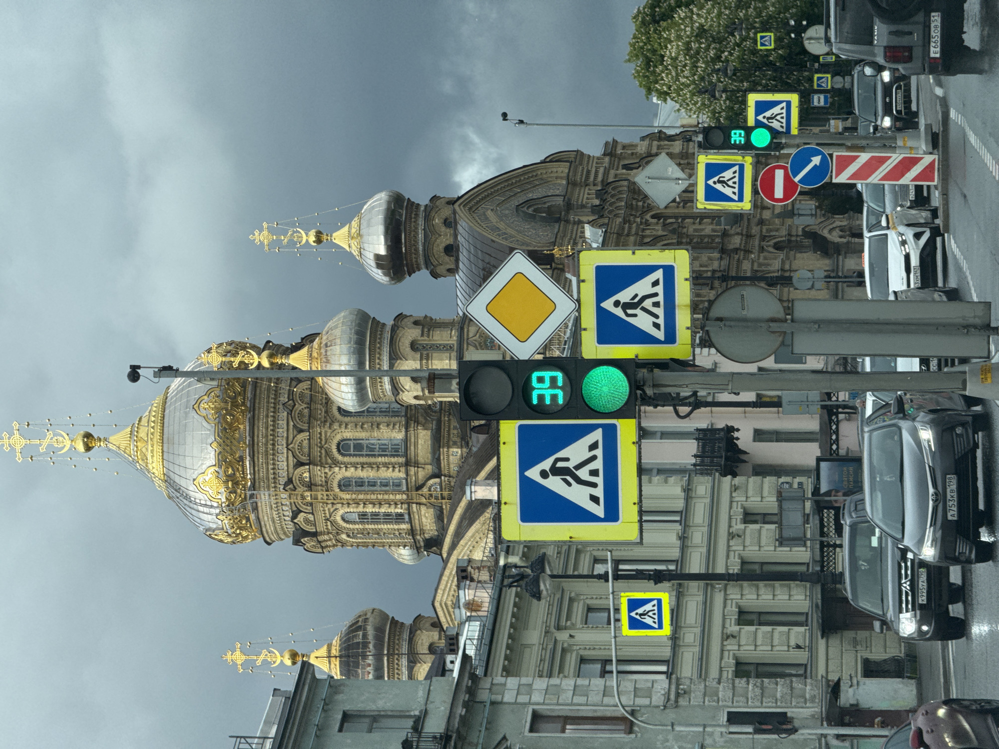 The image captures a street scene in Saint Petersburg, Russia. A prominent architectural feature is the Orthodox cathedral with its gilded domes and ornate facade. In the foreground, a traffic light shows a green signal with a timer indicating 39 seconds. Several pedestrian crossing signs are visible alongside other road signs, including a "no entry" sign and a sign indicating a direction to turn. Cars are lined up on the right side of the frame, with visible license plates indicating Russian registration. The sky is overcast, suggesting potentially inclement weather.