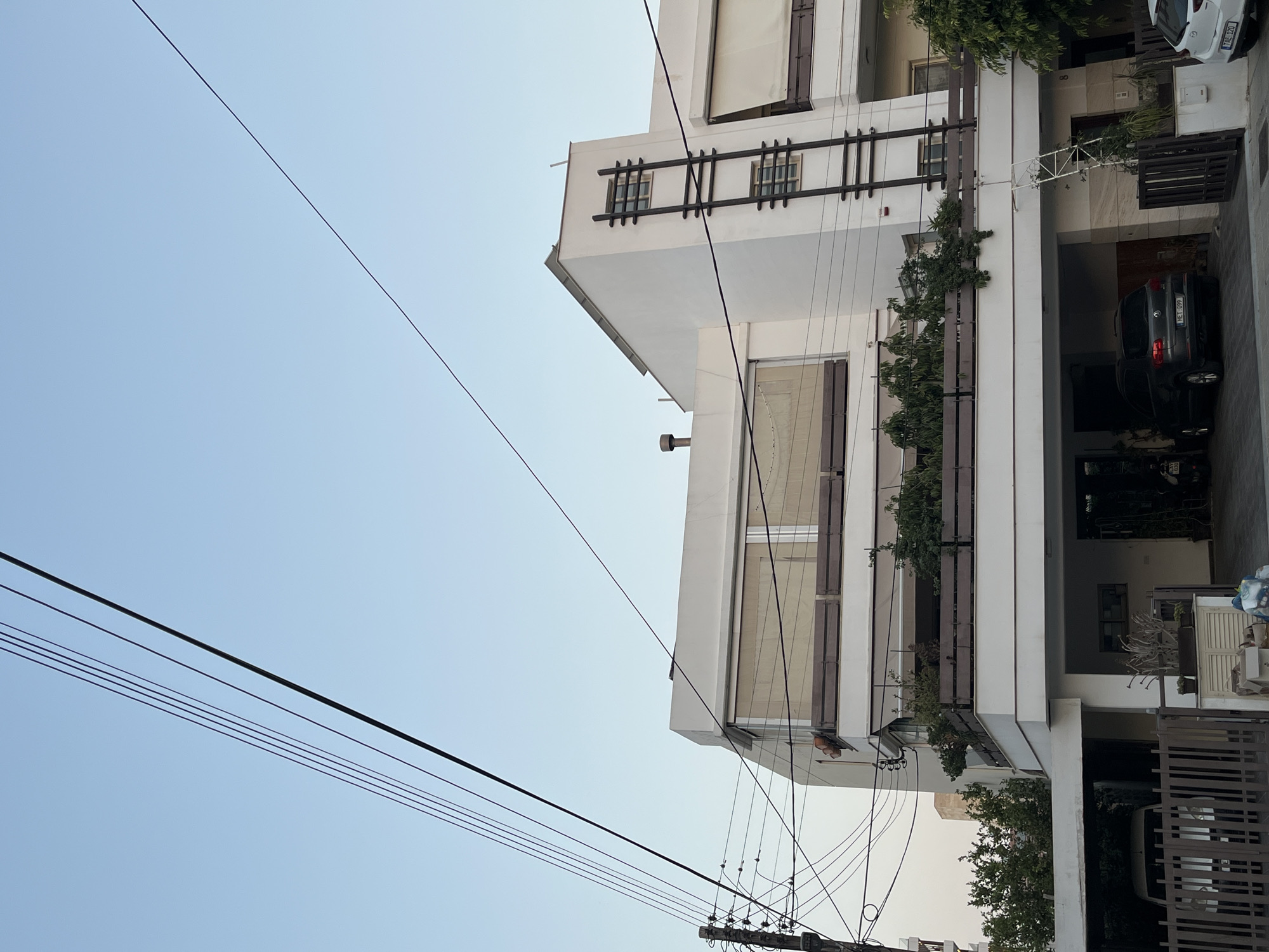 The image depicts a residential building in Limassol, Cyprus. The building is modern in design, with white stucco walls and large windows. A balcony on the second floor is filled with greenery, and an attached carport shelters two vehicles. Overhead, a network of electrical wires stretches across the clear blue sky, obscuring the upper portion of the building. The scene appears to be captured during daylight hours, possibly late afternoon, given the soft lighting. No people are visible in the image, and the overall atmosphere is one of quiet domesticity. The architecture suggests a Mediterranean influence, common in coastal cities like Limassol.