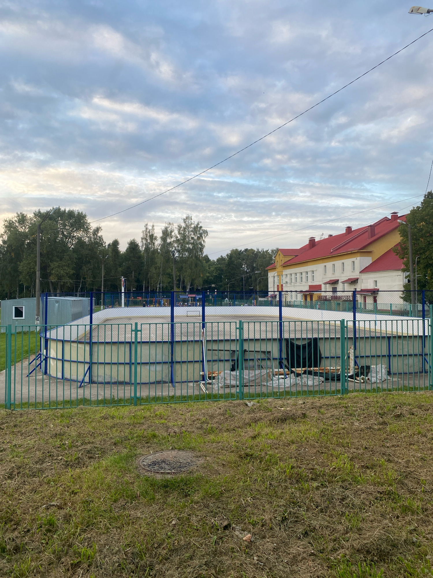 The image depicts an outdoor ice rink under construction in Machulishchy, Belarus. The rink is enclosed by a green metal fence and has a light-colored surface. In the background, a building with a red roof and yellow facade stands behind a line of trees. The sky is overcast with gray clouds. The foreground shows a grassy area with a manhole cover. There are no people visible in the image.