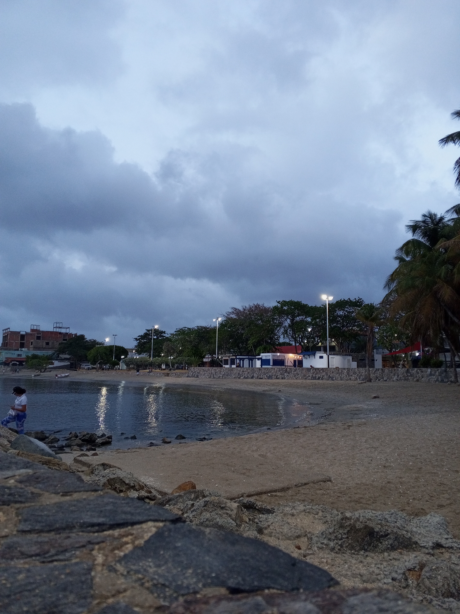 This image captures a coastal scene in Juan Griego, Venezuela, during what appears to be dusk or early night. The dominant feature is a heavily overcast sky filled with dark, formidable grey clouds, creating a moody and subdued atmosphere.
In the foreground, large, dark grey rocks define the immediate edge, transitioning to a sandy beach that stretches along the water. A calm body of water, likely a bay or sea, occupies the mid-ground, beautifully reflecting the bright, vertical streaks of light from several illuminated lampposts lining the shore.
Along the coastline, various structures are visible. On the far left, a person is standing by the water's edge near the rocks, possibly looking down. Further back, an unfinished building with exposed rebar stands prominently, and a small boat is visible near the shore. The middle ground features several small, distinct buildings, some painted white and blue, possibly housing cafes or shops, nestled among a line of lush green trees. Palm trees are distinctly visible on the right side, characteristic of a tropical environment.
The artificial lights scattered along the shore suggest active spaces even under the dark sky. The only visible text is "HOTEL," faintly discernible on a building in the far-left background. The scene conveys a quiet, twilight moment with an air of potential weather change, typical of a coastal town.