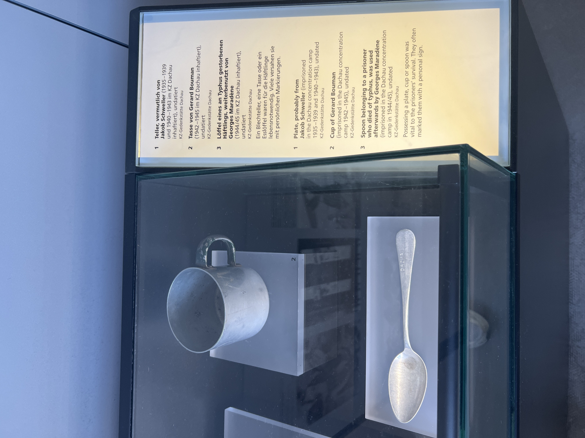 The image shows a display case containing a metal spoon and a metal mug, possibly from a concentration camp. The display is accompanied by a text panel that describes three items. The first item is a plate, likely from Jakob Schweller, who was imprisoned in the Dachau concentration camp from 1935-1939 and 1940-1943. The second item is a cup belonging to Gerard Bouman, who was imprisoned in the Dachau concentration camp from 1942-1945. The third item is a spoon that belonged to a prisoner who died of typhus and was later used by Georges Maradène, who was imprisoned in the Dachau concentration camp in 1944/45. The text further explains that possessing a plate, cup, or spoon was vital for the prisoners' survival, and they often marked these items with personal signs. The spoon in the display case has the word "DACHAU" inscribed on it. The text panel is in German and English. The location is the Dachau Concentration Camp Memorial Site in Dachau, Germany.