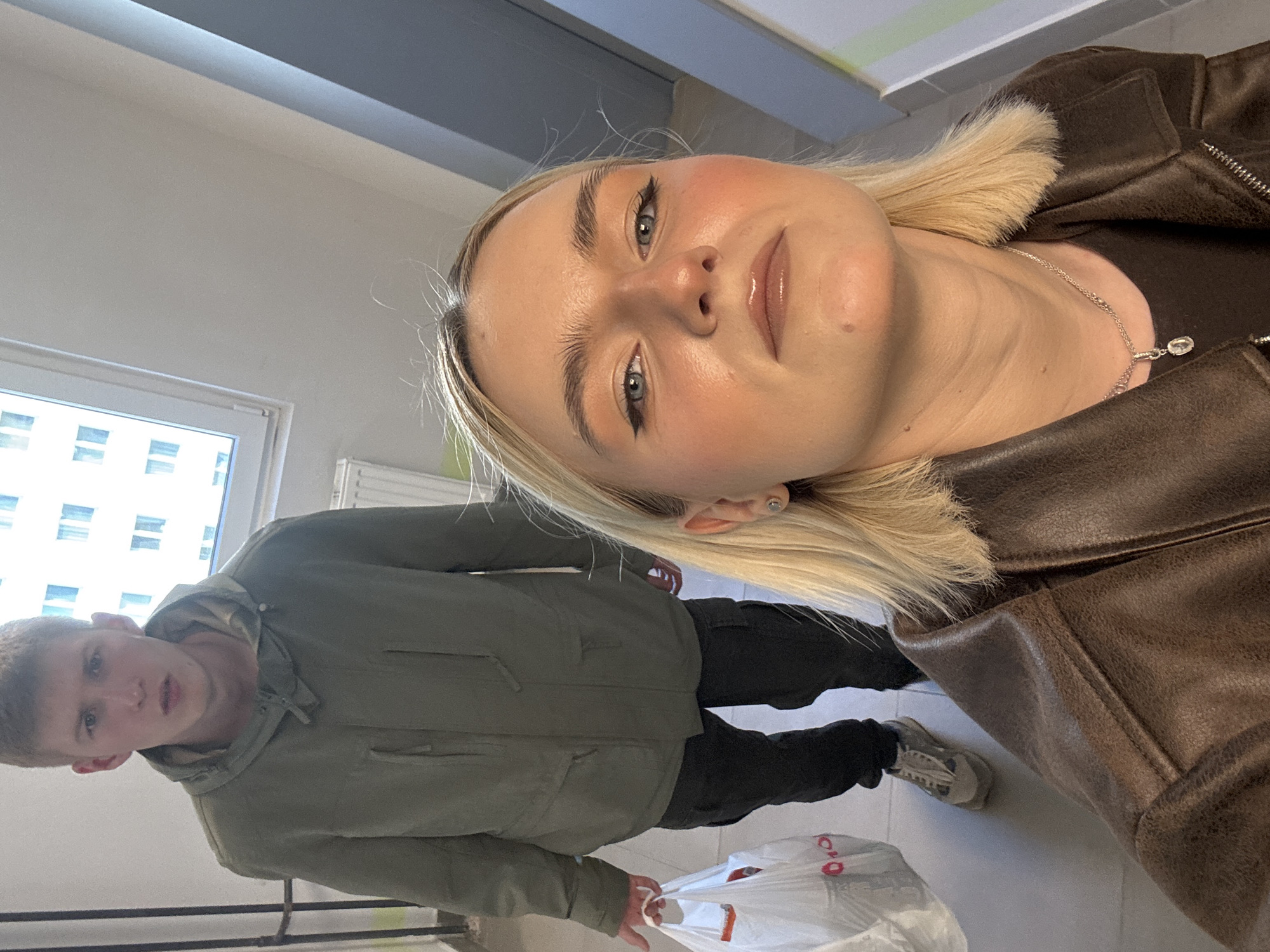 Two individuals are visible indoors. A young woman with blonde hair, wearing a brown leather jacket and a necklace, is in the foreground. A young man, wearing a green jacket and black pants, is in the background holding a white plastic bag. The setting appears to be a building corridor with a window on the left. There is no evidence to indicate the location is in Irinovka, Russia.