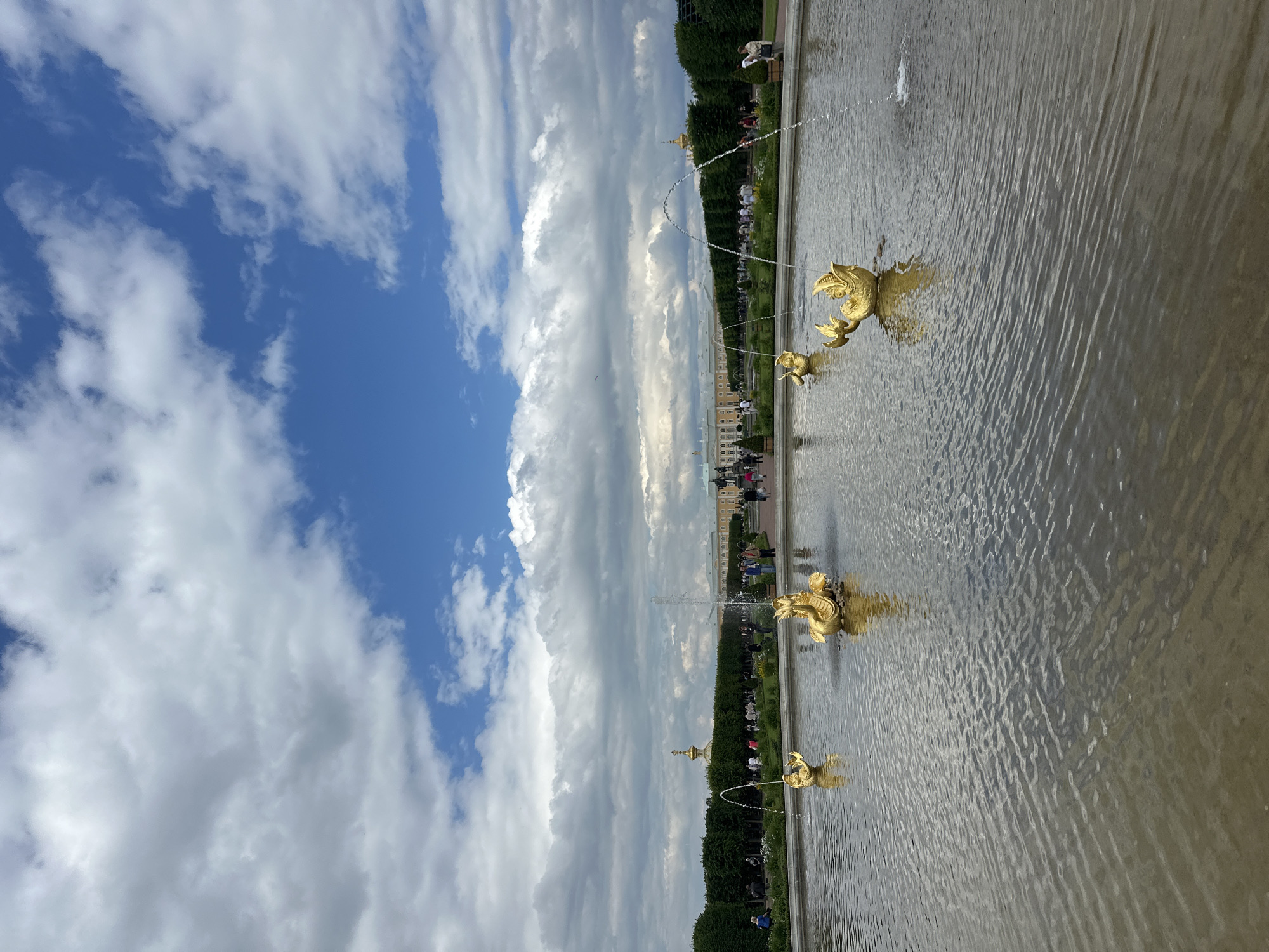 This is a photograph of the Grand Cascade at Peterhof Palace in Peterhof, Russia. The image shows a large fountain in the foreground with several golden sculptures of mythical sea creatures, each spouting water. The fountain is situated in a wide, calm pool of water that reflects the sky and the surrounding greenery.
In the background, a grand palace complex with yellow facades and gilded domes is visible. A line of manicured trees forms a hedge along the perimeter of the palace grounds, and a path runs alongside it, populated by numerous visitors strolling and observing the fountains. The sky above is a mix of bright blue with dramatic, large white clouds. The overall impression is one of an ornate and impressive Baroque garden.
