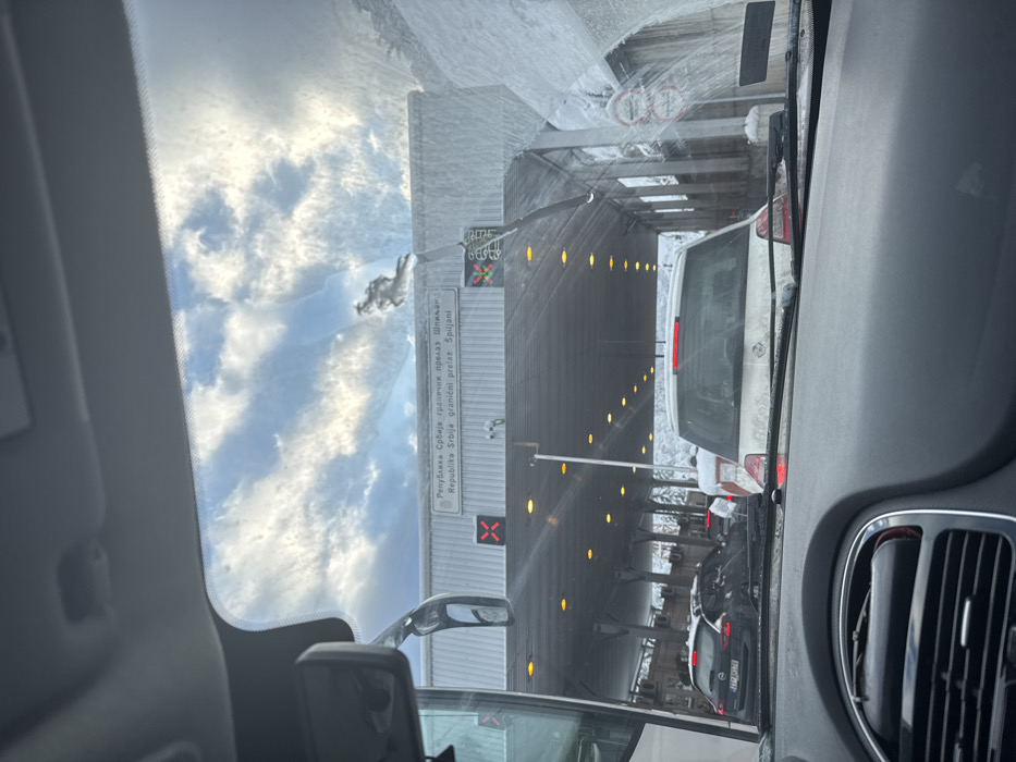 A frontal view from inside a vehicle captures the Špiljani border crossing in Tutin Municipality, Serbia, under snowy, daytime conditions. The upper section of the vehicle's dashboard, rearview mirror, and a streaked windshield are visible in the foreground. Directly ahead, a multi-lane border crossing structure, heavily covered in snow, displays the sign "Republika Srbija granični prelaz Špiljani" in both Cyrillic and Latin scripts. Beneath the structure, multiple lanes are illuminated by amber overhead lights, with red 'X' indicators visible above some lanes, suggesting a stop or closed status. A light-colored hatchback is stationary immediately in front of the observation vehicle, and a dark-colored Opel hatchback with a Serbian license plate is visible in an adjacent lane further forward. Additional vehicles are queued in the distance. Snow accumulates on all visible surfaces, including vehicles and the border infrastructure, beneath a partly cloudy sky. No discernible individuals or active interactions are depicted.