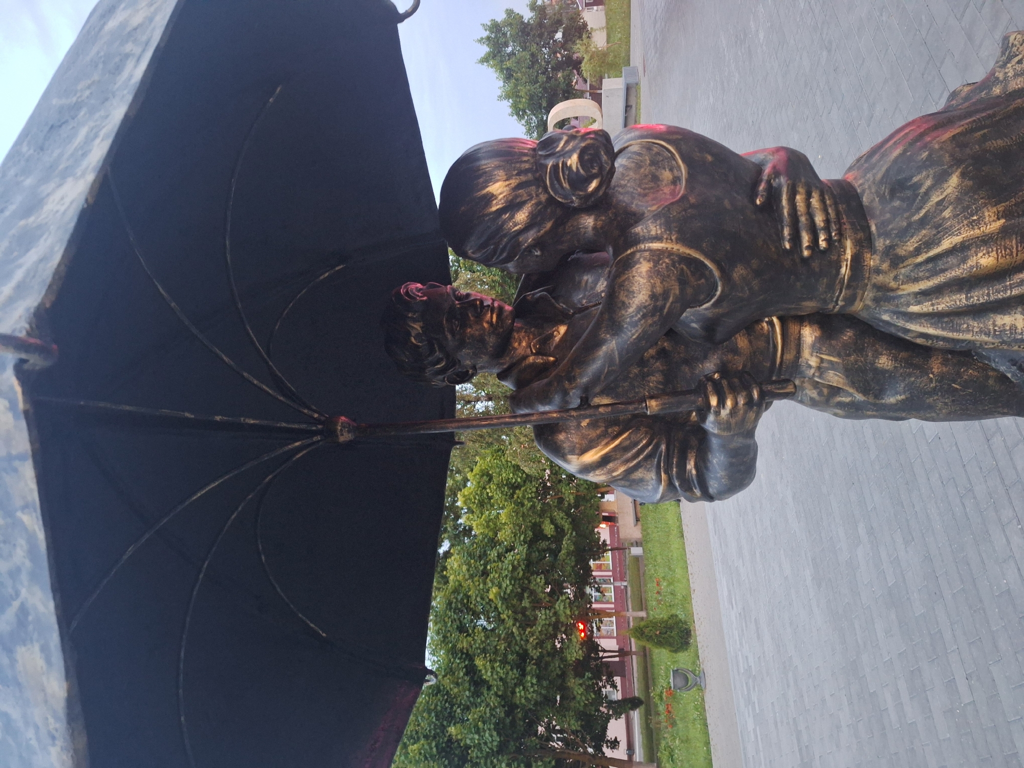 This is a medium-shot, eye-level, outdoor photograph of a bronze sculpture depicting a couple embracing under a large, dark umbrella. The sculpture is rendered in a dark bronze with gold accents that highlight the figures' clothing and features. The male figure is on the left, with his arms around the female figure. The female figure is on the right, facing away from the camera, with her arms around the male figure's neck. They appear to be dancing. The umbrella is held by the male figure, with its spokes visible from underneath.
The sculpture is situated on a paved area made of rectangular, grey bricks. In the background, there is a grassy area with scattered trees and some red flowers. A small, conical evergreen tree is visible to the left of the sculpture, and a grey trash receptacle is to its left. Behind the grassy area, there is a building with a light pink facade and a red roof, and further in the background, more trees are visible against a hazy sky. The setting suggests an urban park or public space in the city of Kapyĺ, Belarus. The lighting is dim, possibly indicating early morning or late evening.