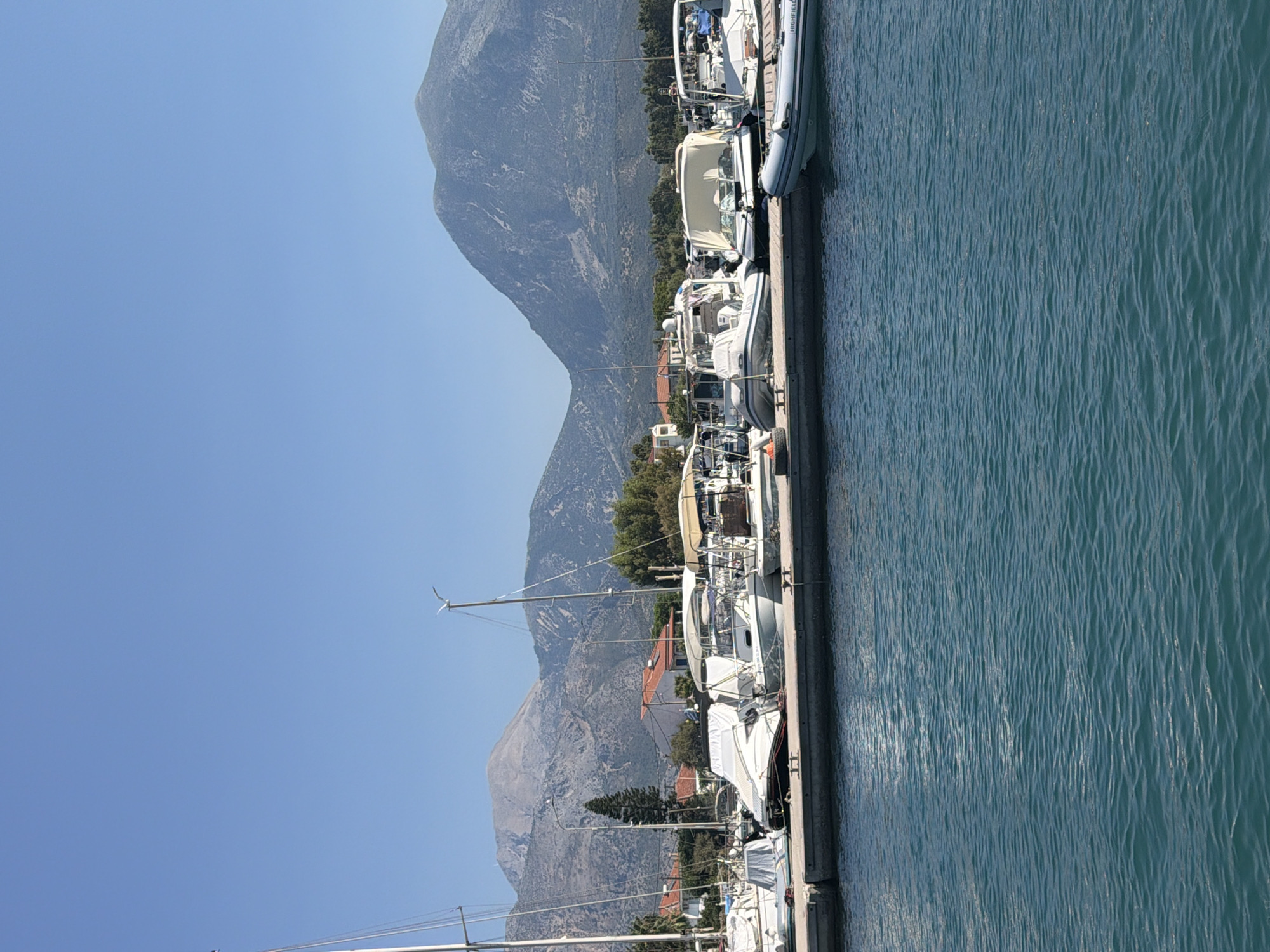 A wide-angle, eye-level, outdoor photograph captures a harbor scene under a clear blue sky. The foreground is dominated by the turquoise water of the harbor, with gentle ripples disturbing its surface. A concrete dock extends horizontally across the midground, where several boats are moored.
On the dock, a collection of boats of various sizes and types are visible. To the far left, the mast and rigging of a sailboat are prominent, with its hull covered by a white tarp. Next to it are several other motorboats and small yachts, some with their covers on, others more exposed. One boat has a beige canopy providing shade. A white and grey inflatable dinghy with "HIGHFIELD" visible on its side is positioned in the water to the right of the dock.
In the background, beyond the dock and boats, is a coastal town with buildings featuring orange-tiled roofs. Lush green trees and foliage are scattered throughout the town and along the hillsides. Towering in the far background are majestic, rugged mountains, their slopes textured with sparse vegetation and rocky outcrops. The overall impression is one of a serene and picturesque maritime setting in the Municipal Unit of Alyzia, Greece.