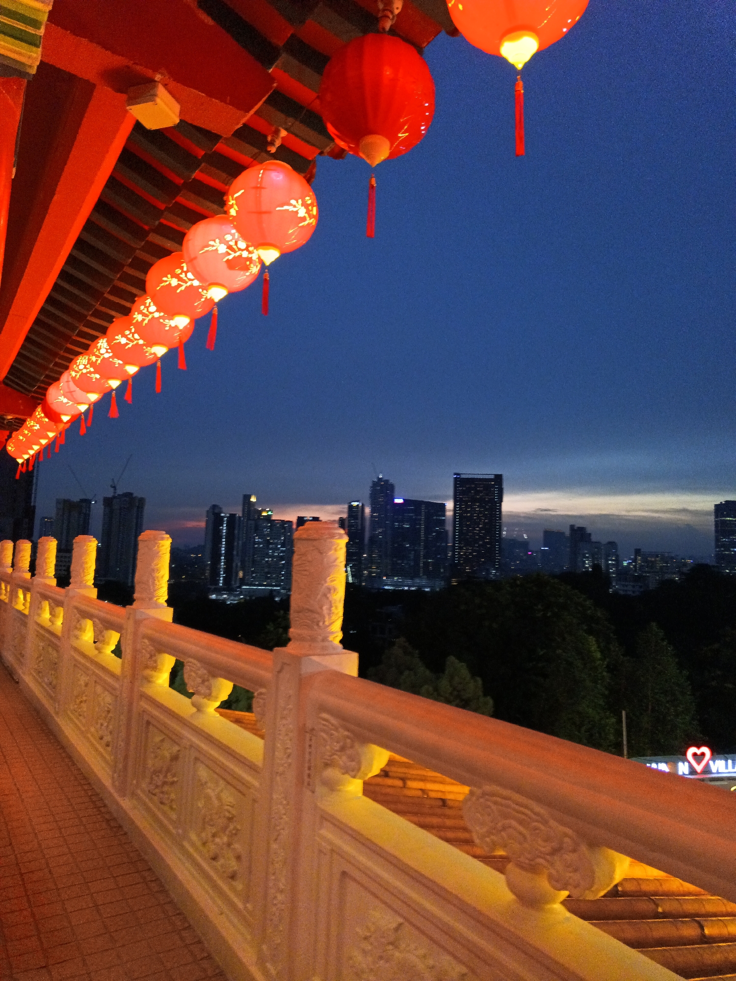 The image depicts a view from a balcony or walkway adorned with ornate white balustrades and red-tiled roofing, illuminated by warm yellow lights. A string of red lanterns, some with decorative Chinese characters, hangs from the eaves, casting a festive glow against the twilight sky. In the background, the Kuala Lumpur cityscape unfolds, showcasing a collection of modern skyscrapers illuminated at dusk. The sky transitions from deep blue to a softer hue near the horizon, suggesting the end of daylight. The scene appears to be from a vantage point in Kuala Lumpur, possibly from a temple or a building with a view of the city's iconic skyline. There are no people visible in the frame.
