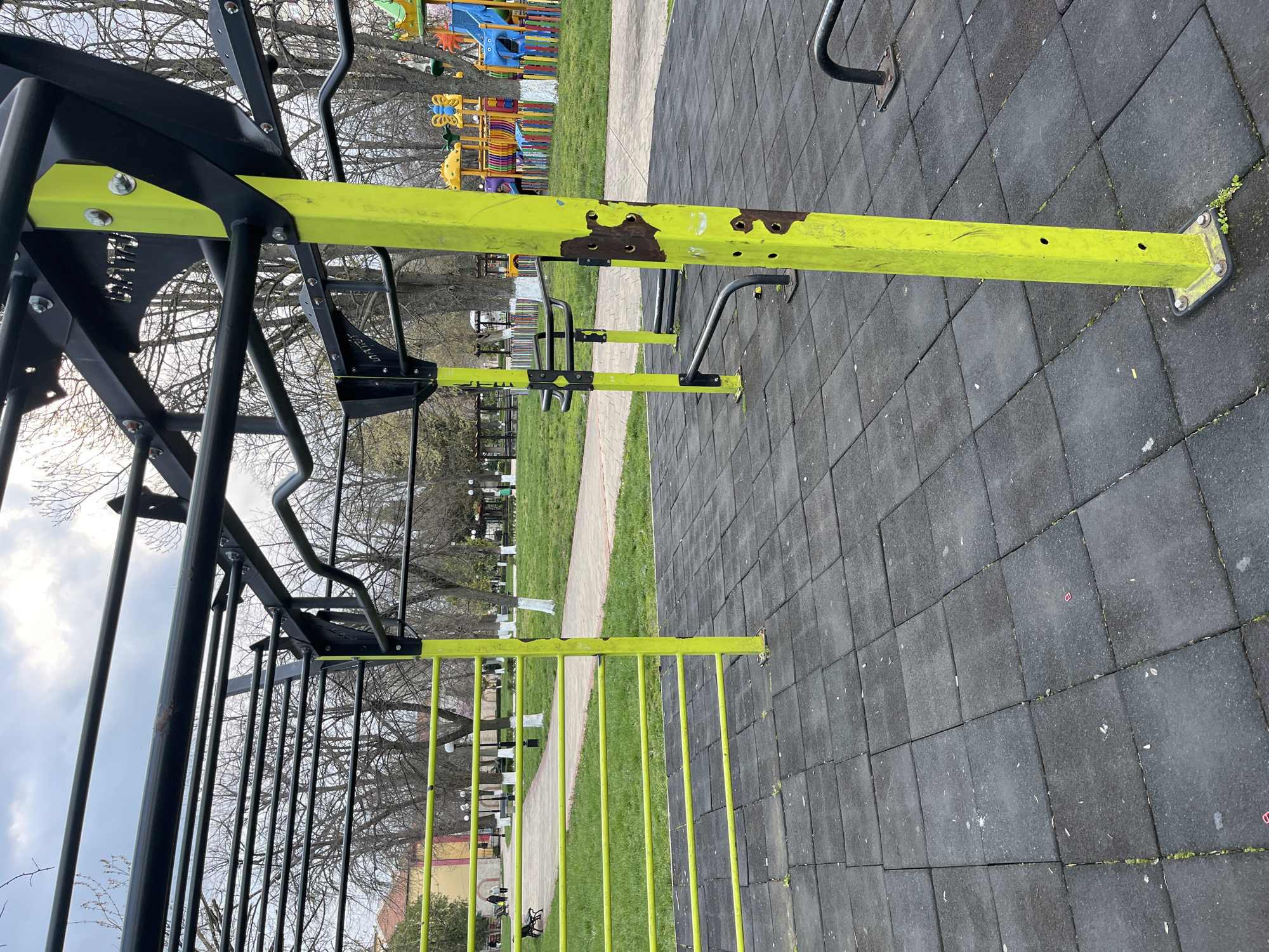 The image shows a playground or outdoor fitness area in Nova Zagora, Bulgaria. It features a set of monkey bars and other exercise equipment made of black and yellow metal. The equipment appears to be slightly damaged, with rust visible on some parts. The ground beneath the equipment is covered in gray rubber safety tiles. In the background, there is a grassy area with trees and other playground equipment, including colorful slides and climbing structures. The sky is overcast. There are no people visible in the immediate foreground.